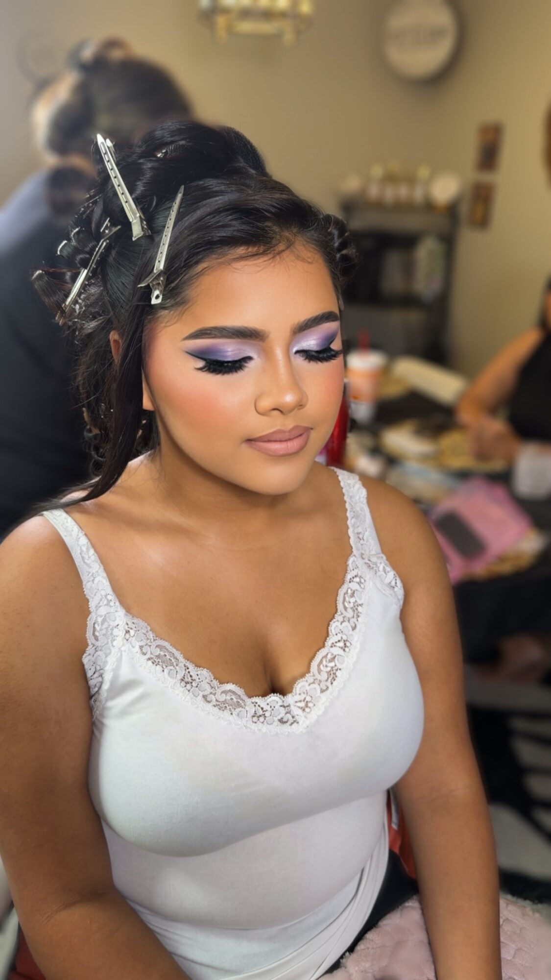 Young woman with makeup and hair styled, wearing a white top with lace, in a room with people and makeup tools