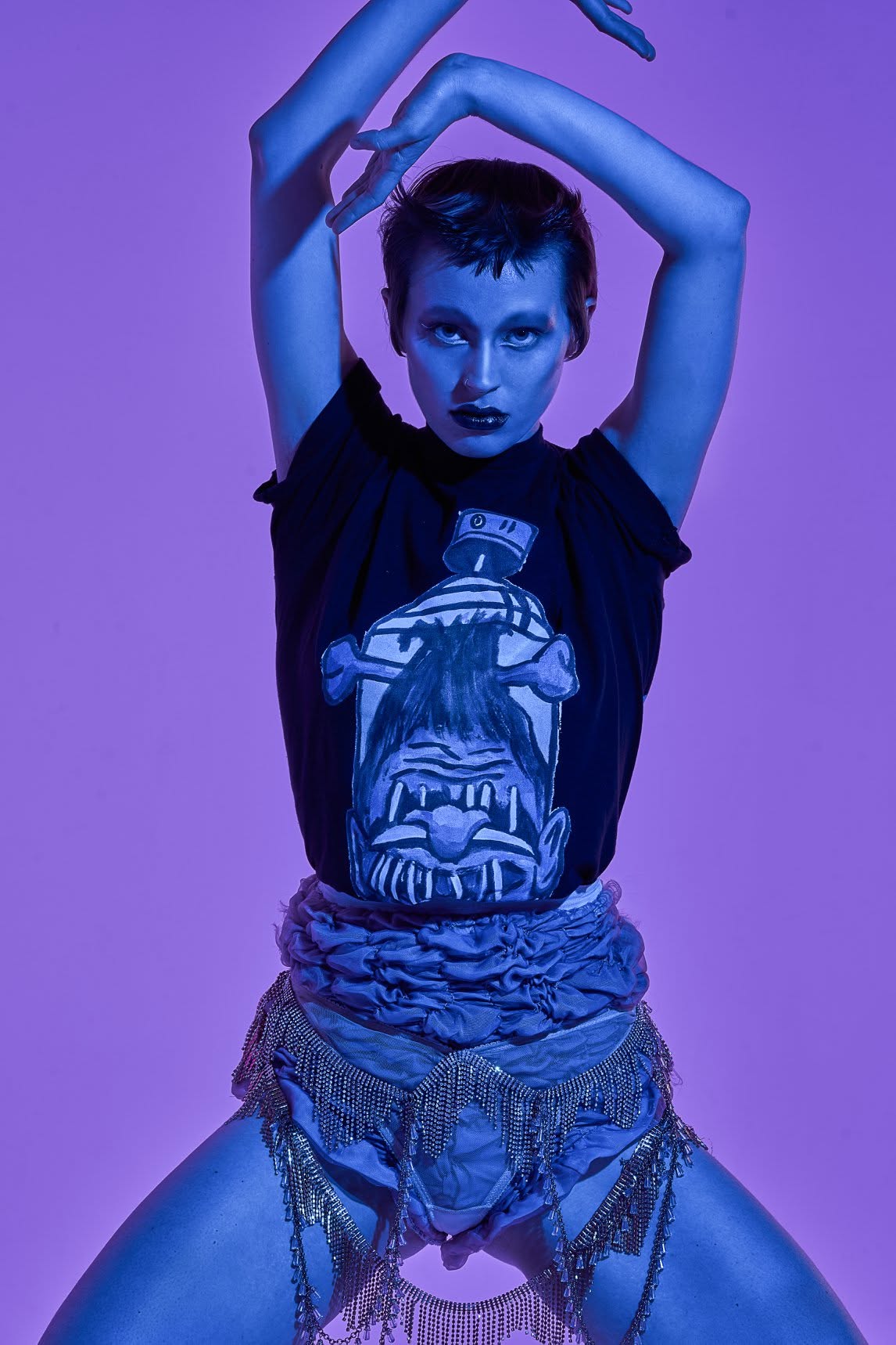 Person with short hair and dark makeup poses with arms above head, wearing a graphic t-shirt and layered accessories, purple background.