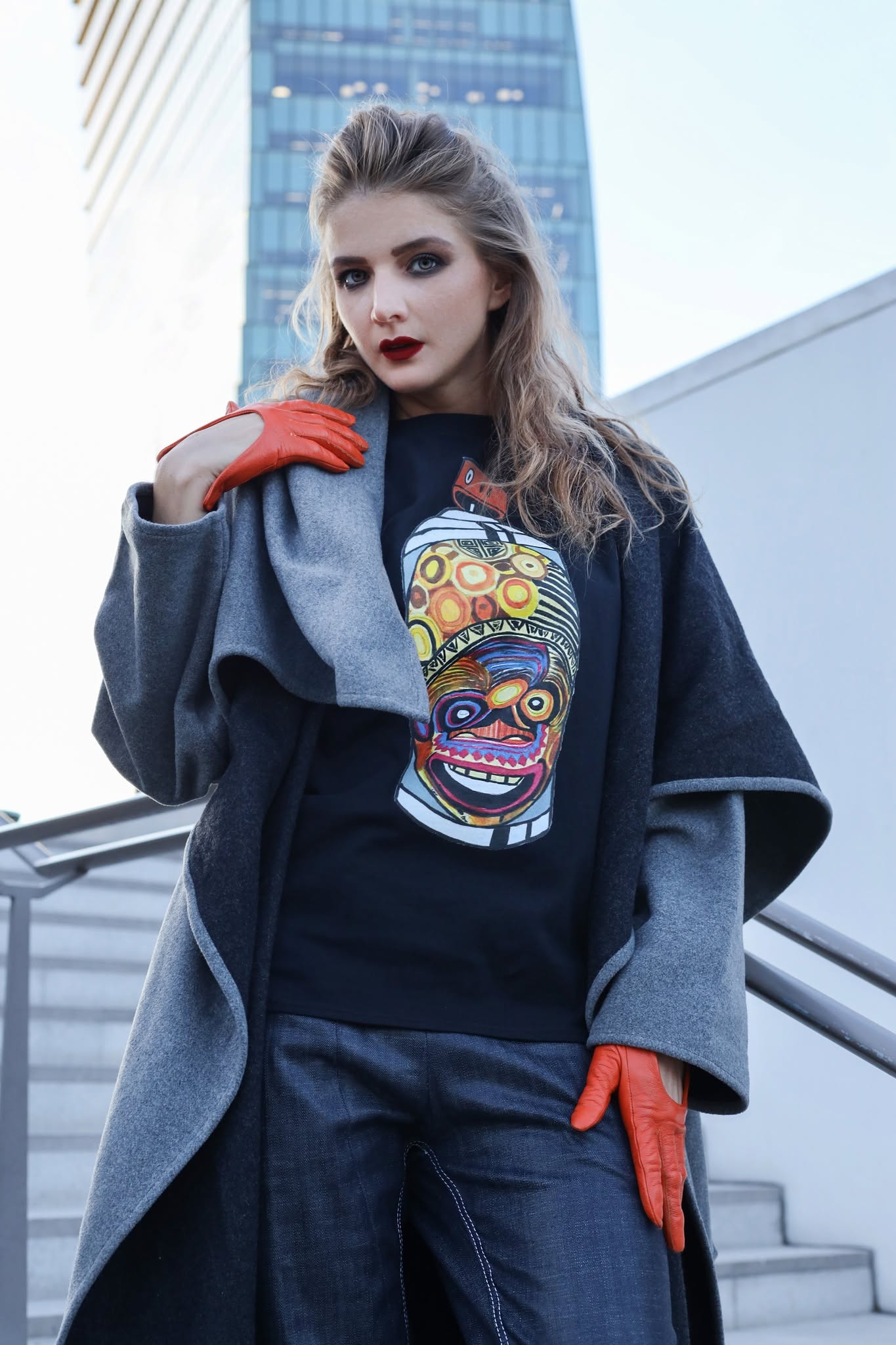 Young woman with wavy hair and dark lipstick stands outdoors in urban setting, wearing a graphic hoodie and orange gloves.