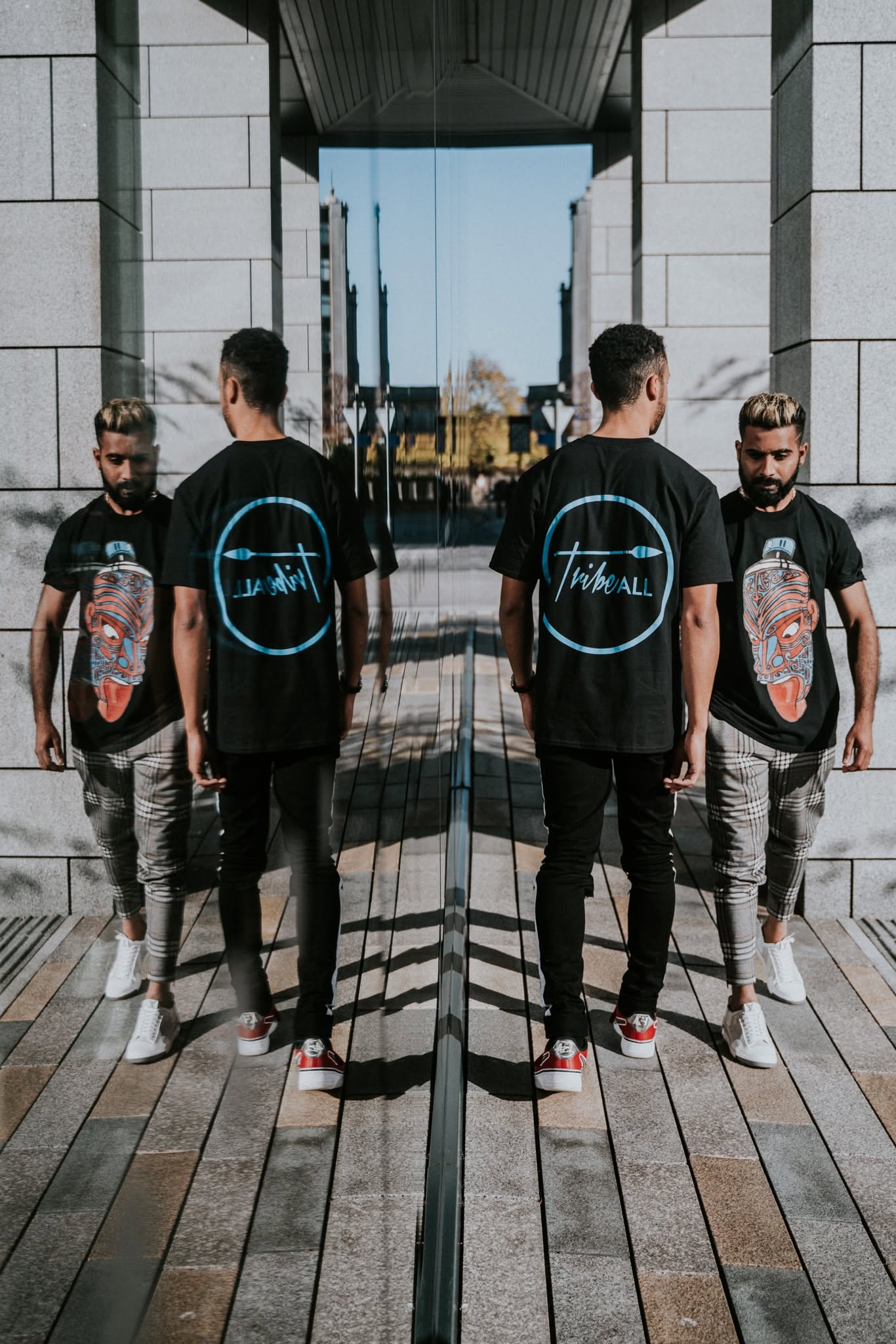 Two men stand in front of a mirror reflecting their images, with a building and sky in the background.