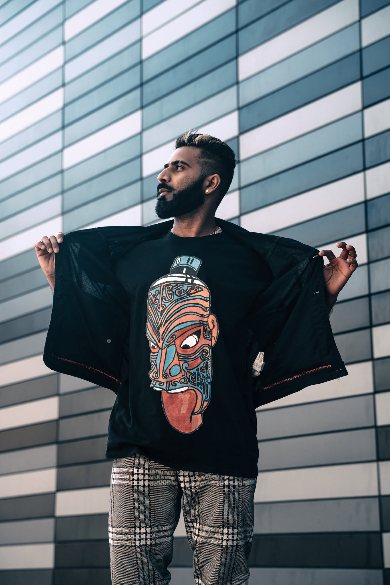 Man with beard and glasses stands in front of a modern glass building, holding open his jacket to reveal a graphic T-shirt.