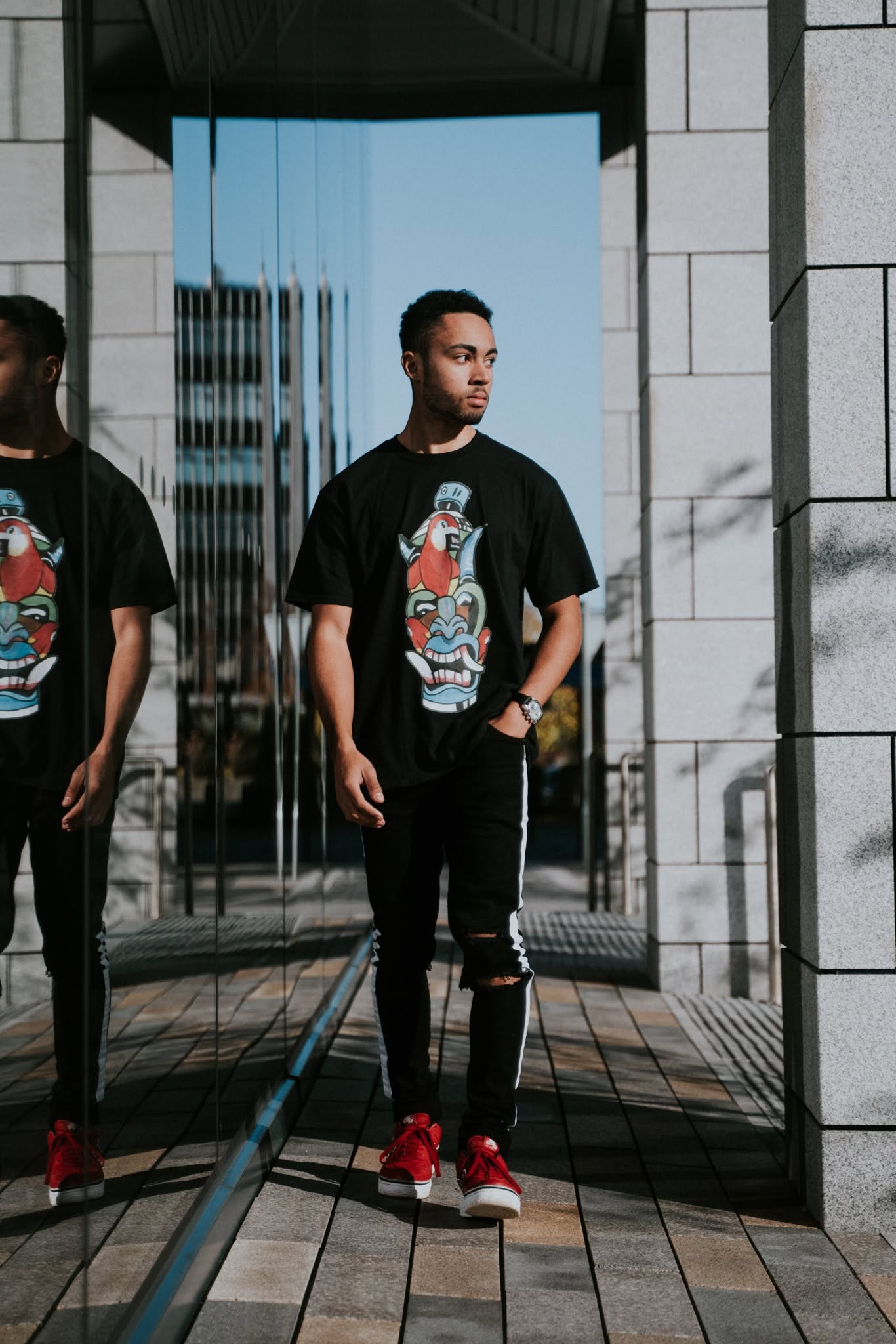 Young man standing outdoors in front of reflective glass and stone building, wearing a black graphic t-shirt, black pants, and red shoes.