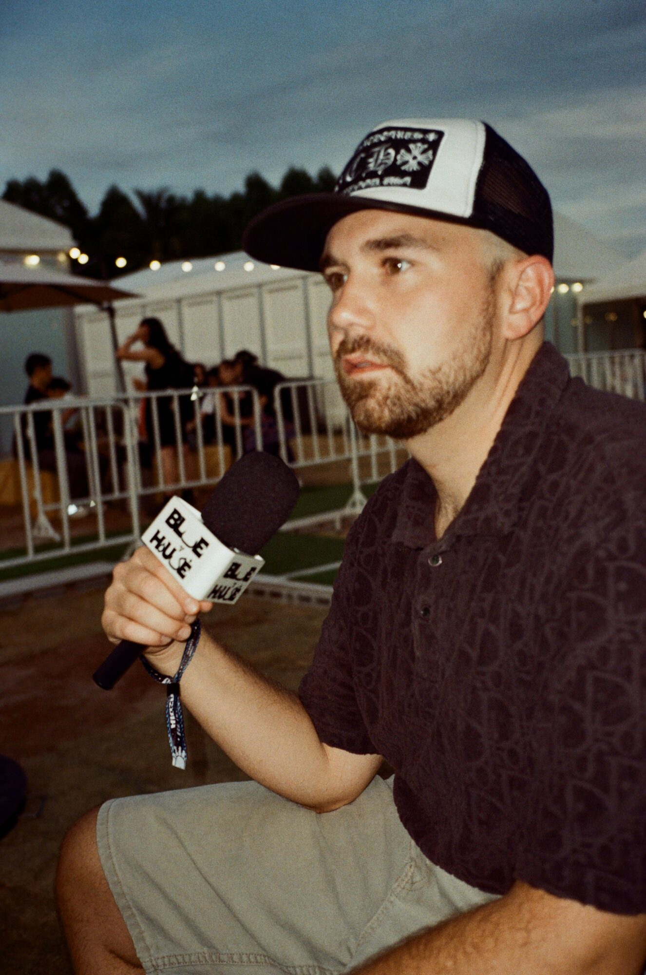 Man wearing a black shirt and a white cap holding a microphone outdoors during evening, with a fence and people in background.
