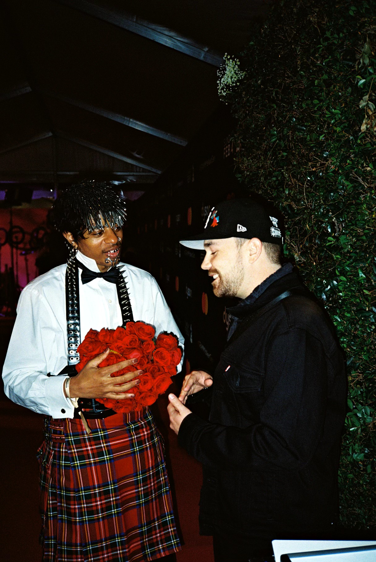 Two men talking, one holding a bouquet of red roses, in a dimly lit setting with greenery background.