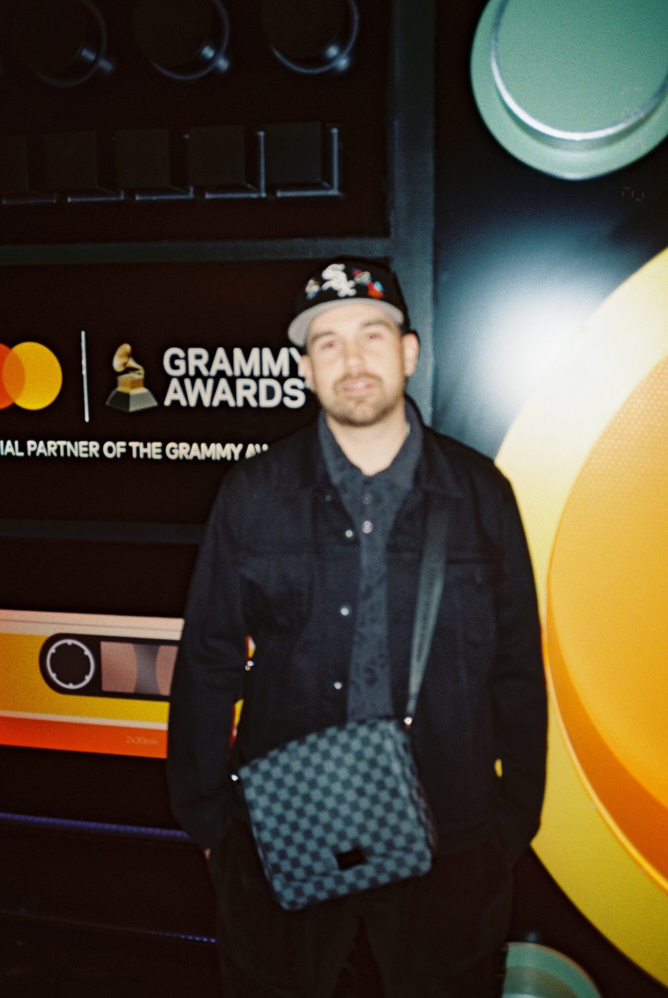 Man wearing a black jacket and cap, carrying a checkered bag, standing in front of Grammy Awards display.