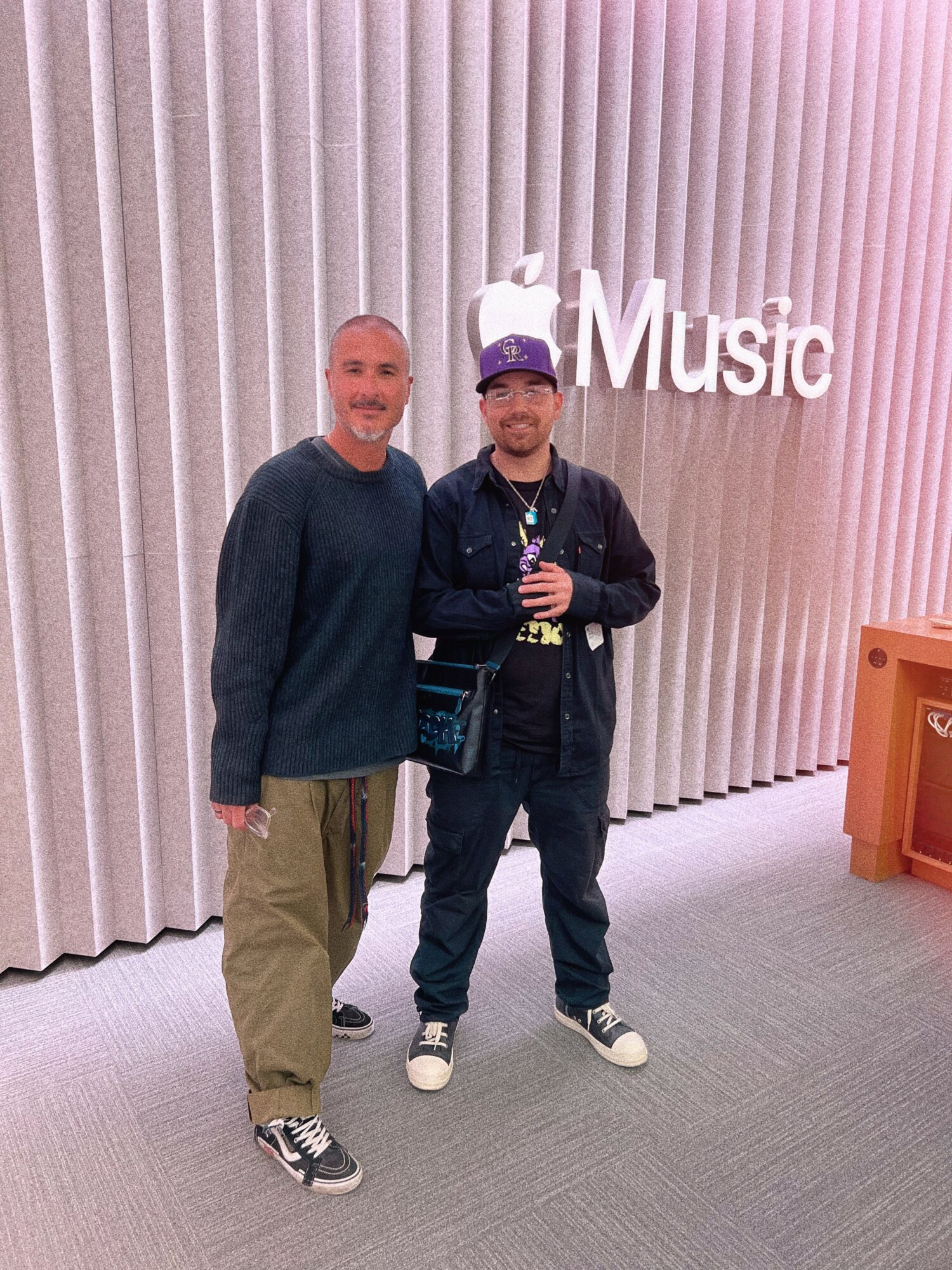 Two men standing in front of a wall with vertical lines and a sign that says 'Music'.