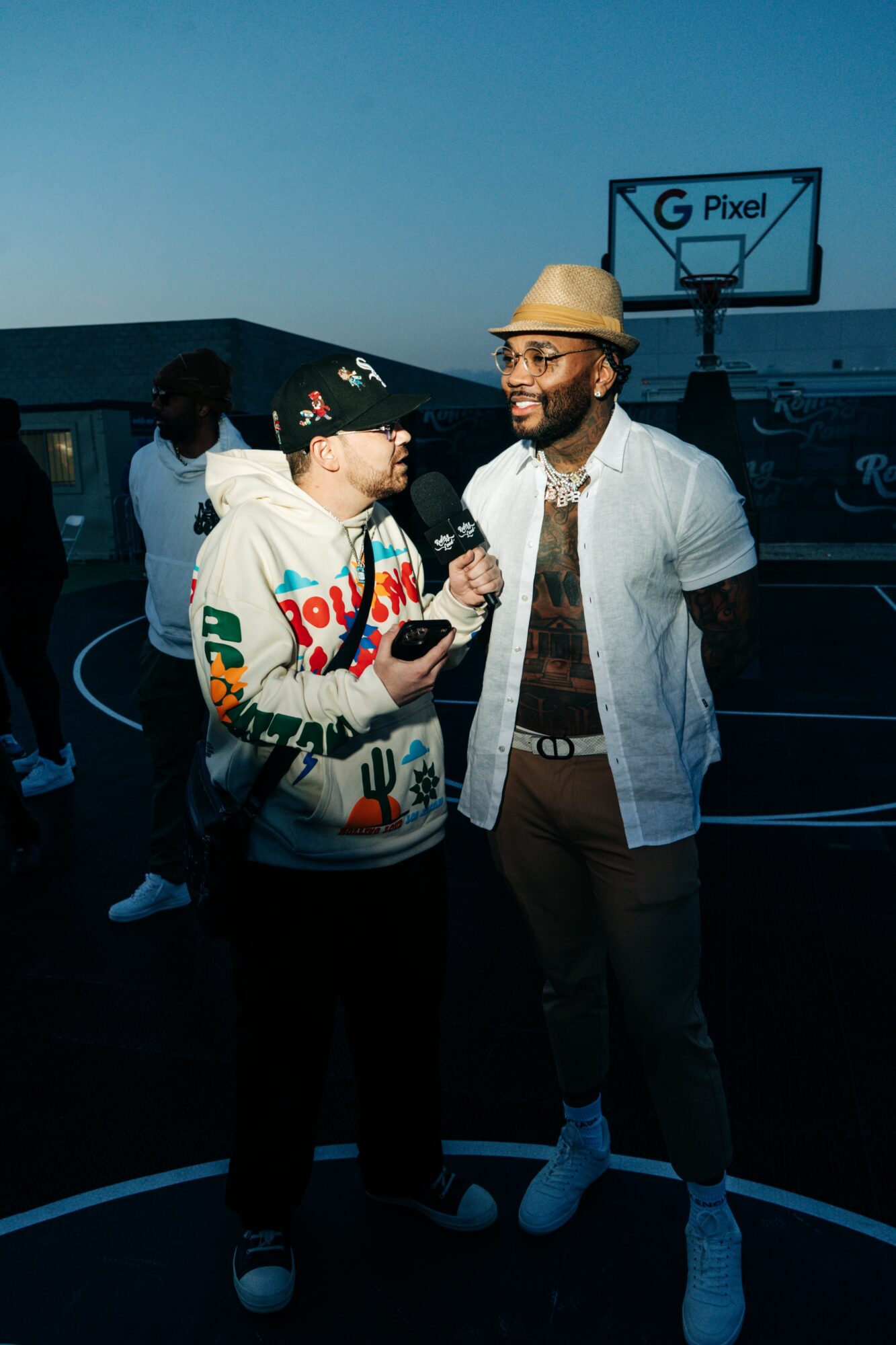 Two men talking outdoors at dusk, one holding a microphone, basketball hoop in background, dark sky, casual clothing