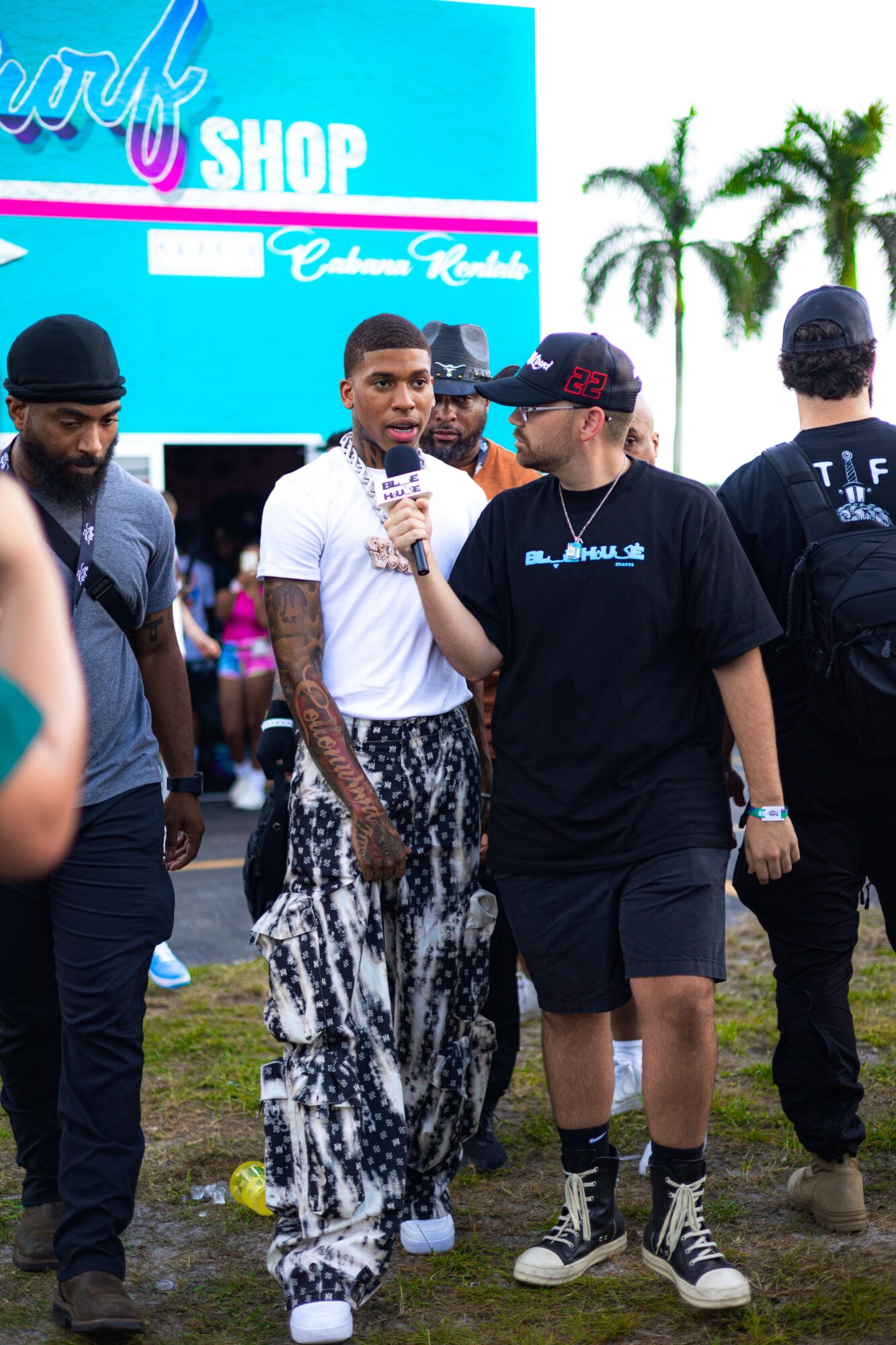 Group of people outdoors near a blue sign with palm trees in background, one person speaking into a microphone, others listening.
