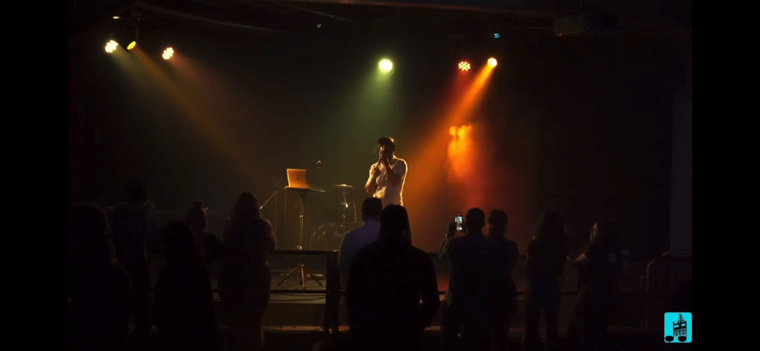 Performer on stage with colorful lighting, audience silhouettes in front, dark background.