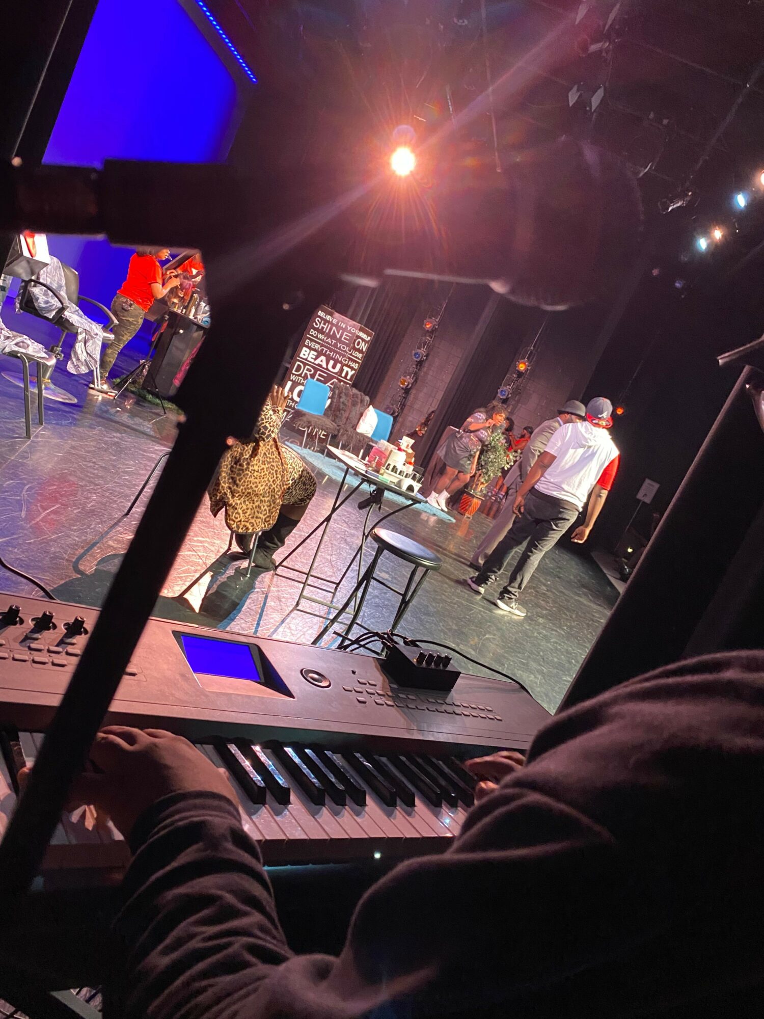 Person playing keyboard on stage with performers and stage lights in background.
