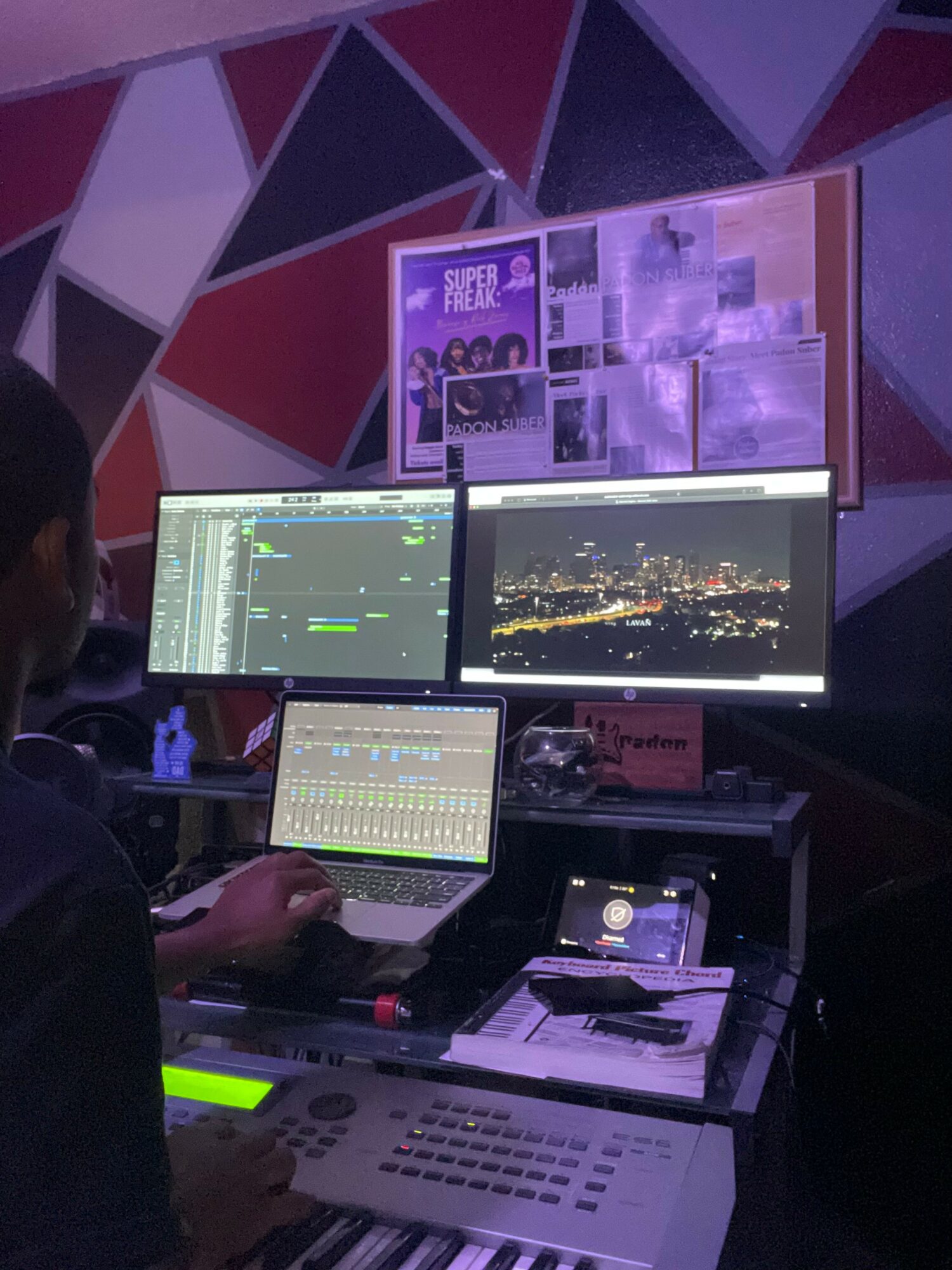 Person working at a control station with three monitors displaying cityscape, editing software, and images, in a dimly lit room.