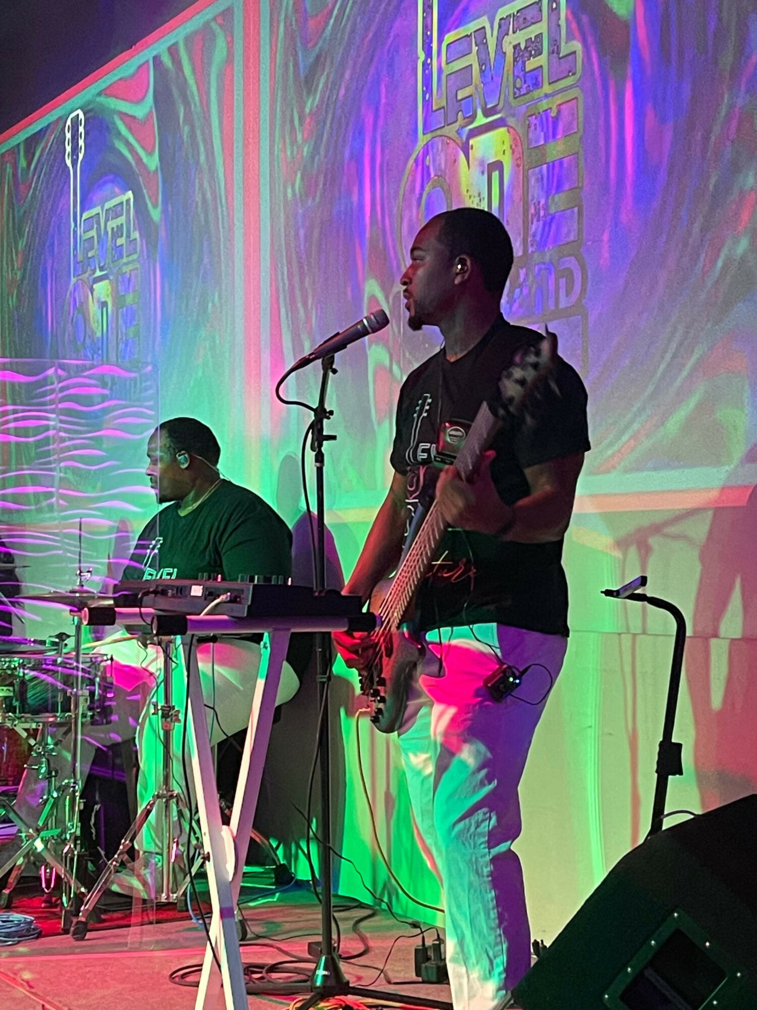 Two musicians perform on stage with colorful lighting and a backdrop, one playing guitar and the other on keyboard.