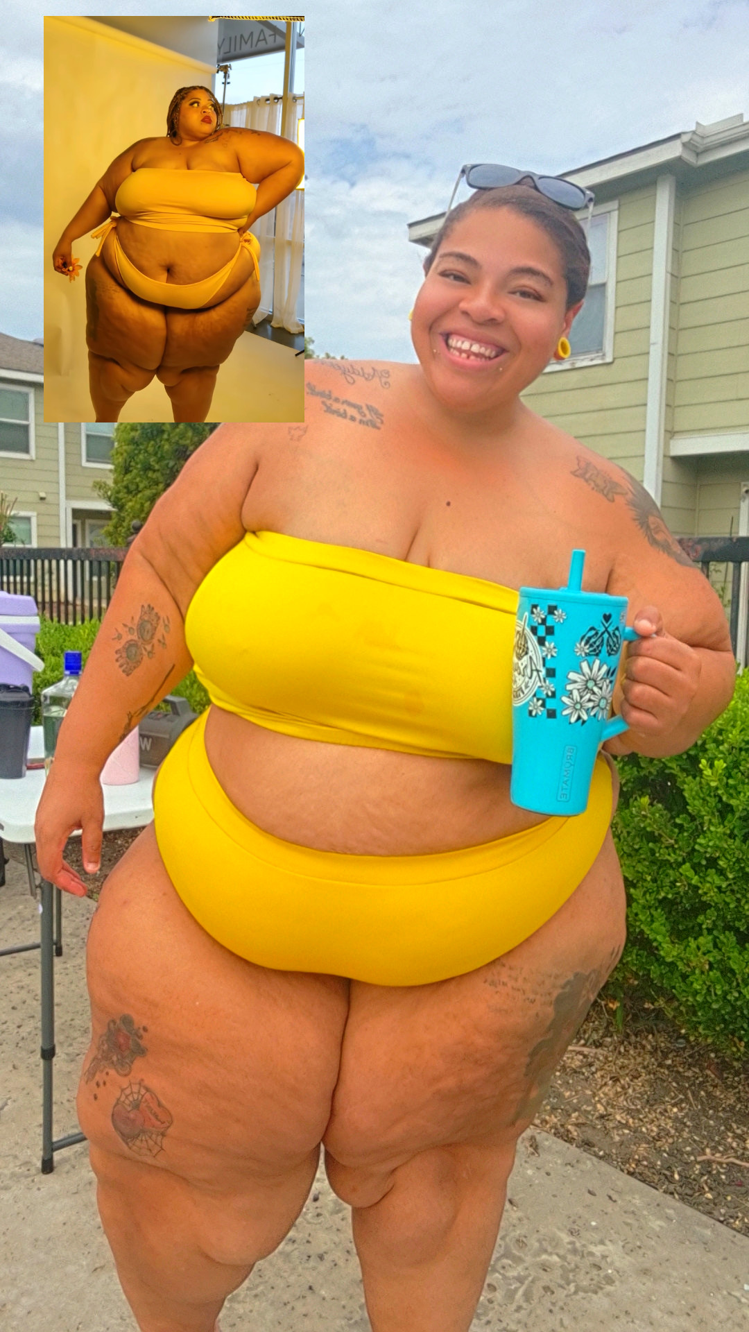 Smiling woman in yellow swimsuit holding a blue cup outdoors, with a house and greenery in background.