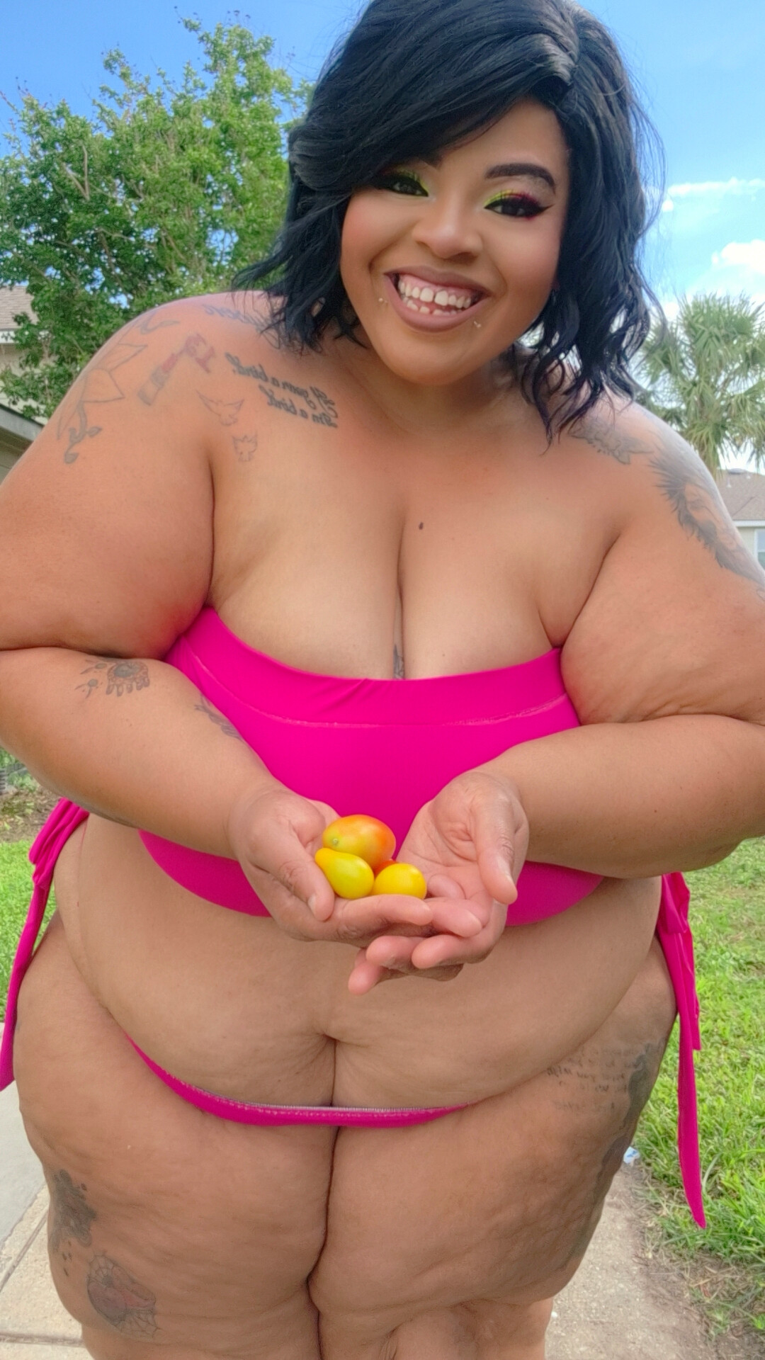 Smiling woman outdoors holding yellow eggs, wearing a pink top and matching bottoms, with trees and sky in background.