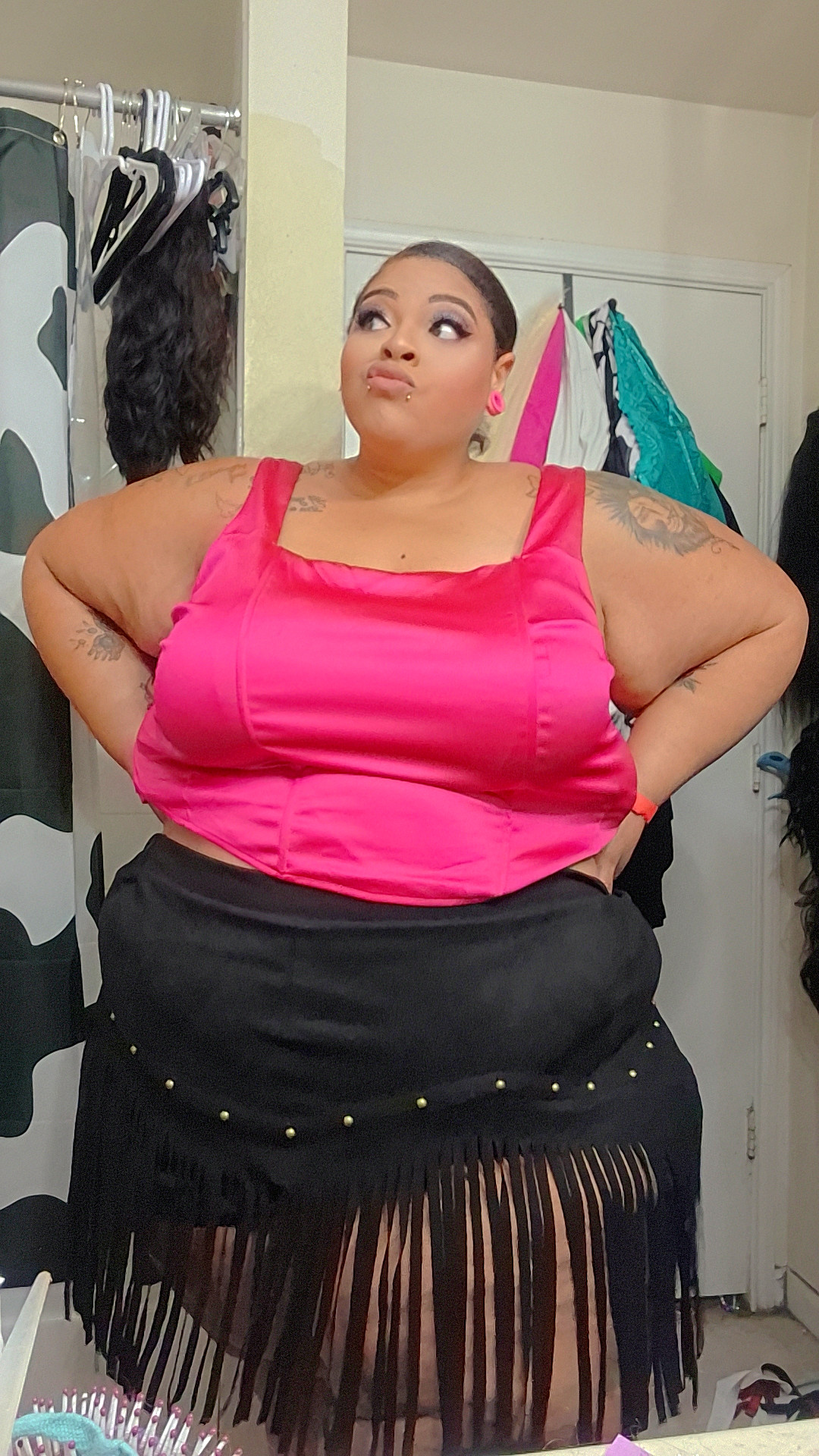 Woman in pink top standing with hands on hips in a room with clothing and accessories, facing forward.
