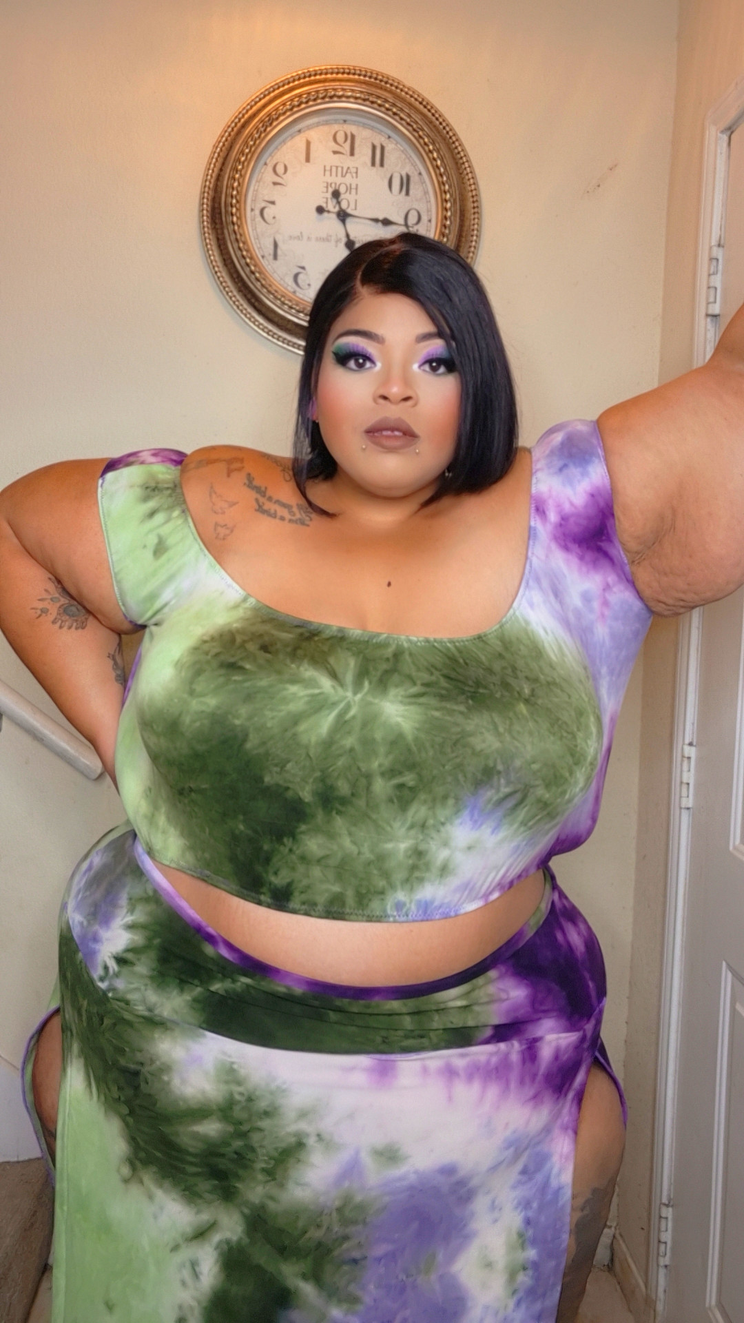 Woman with short black hair and makeup, wearing a green and purple tie-dye outfit, standing indoors near a clock.