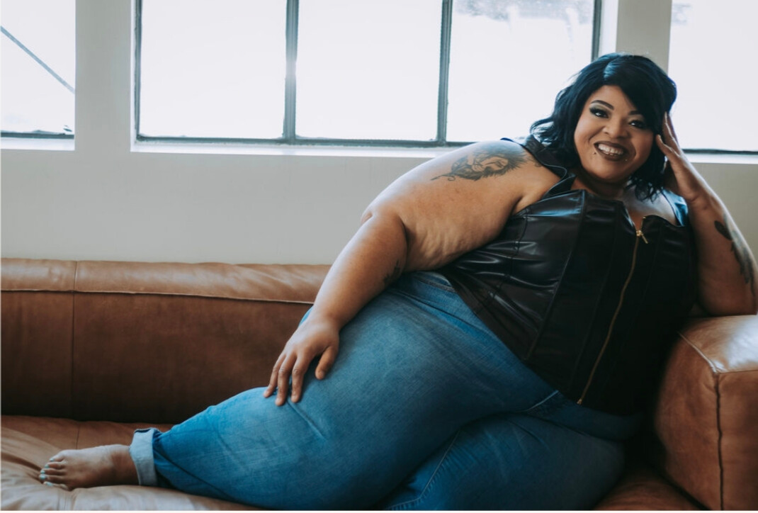 Woman with dark hair and tattoos relaxing on a brown sofa, smiling, in front of windows with natural light.