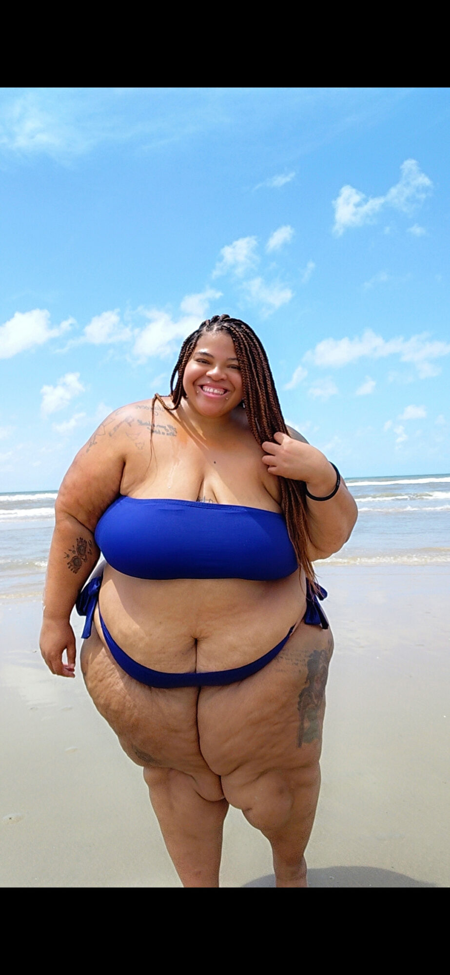 Smiling woman in blue swimsuit on beach with blue sky and clouds, holding her hair, standing on sand.