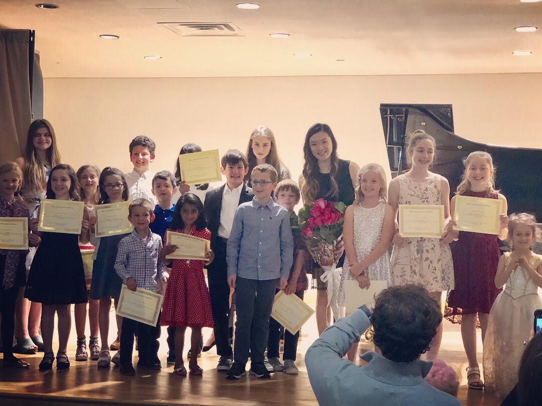 Group of children and adults on stage holding certificates and flowers, with a grand piano in the background.