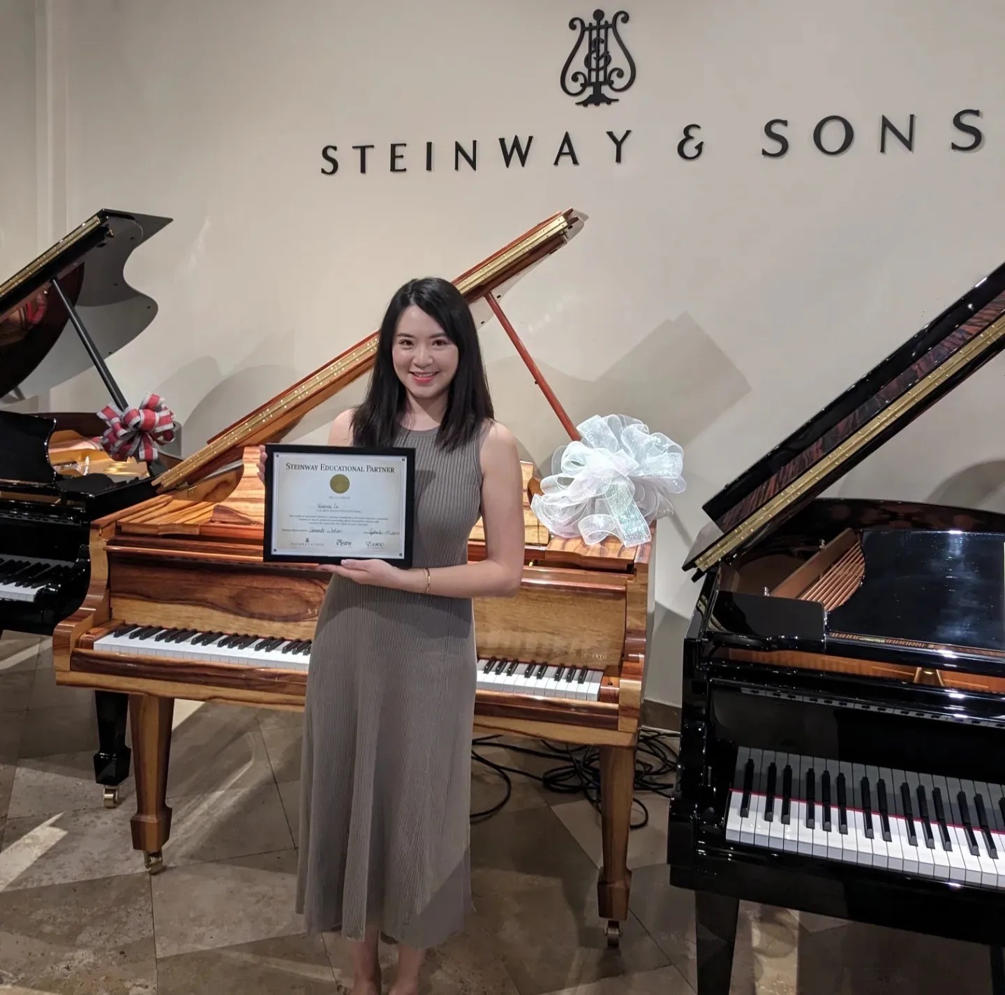 Woman holding a certificate in front of a grand piano, with another piano nearby, in a music store or showroom.