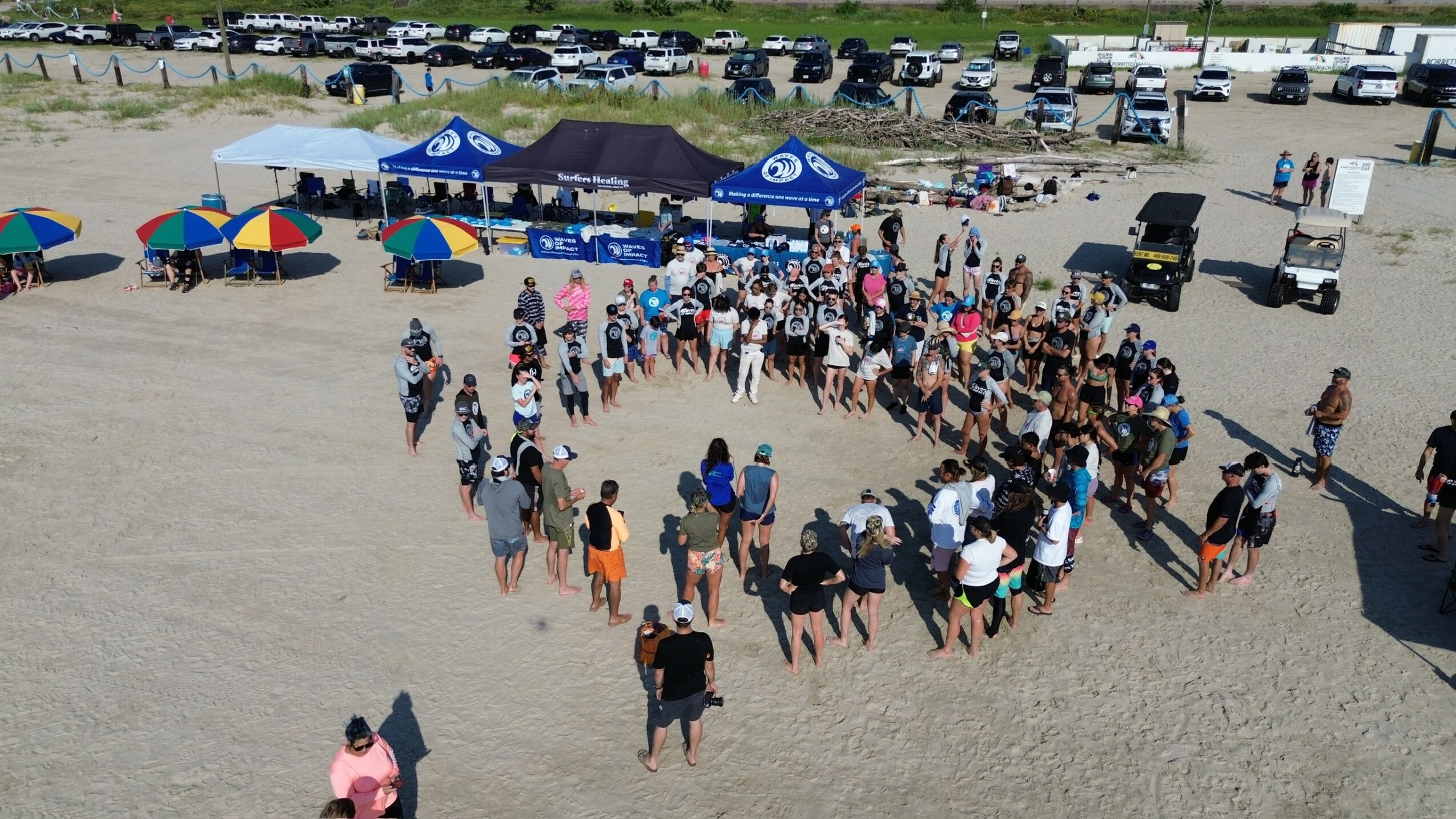 Group of people gathered on a sandy beach, some under tents and umbrellas, with parked cars in the background.
