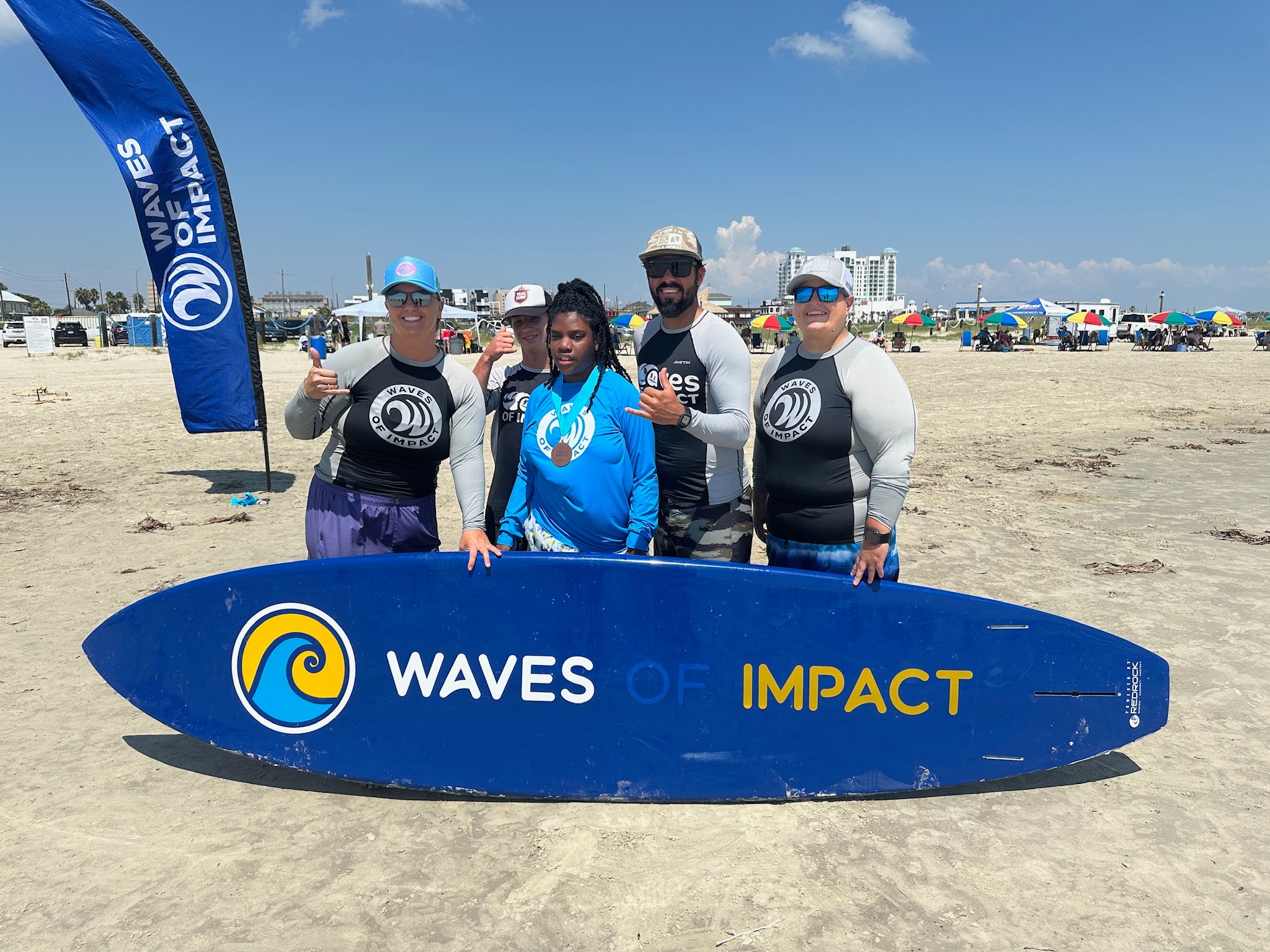 Four people standing on a beach holding a surfboard with 'WAVES OF IMPACT' written on it, blue sky and umbrellas in background.