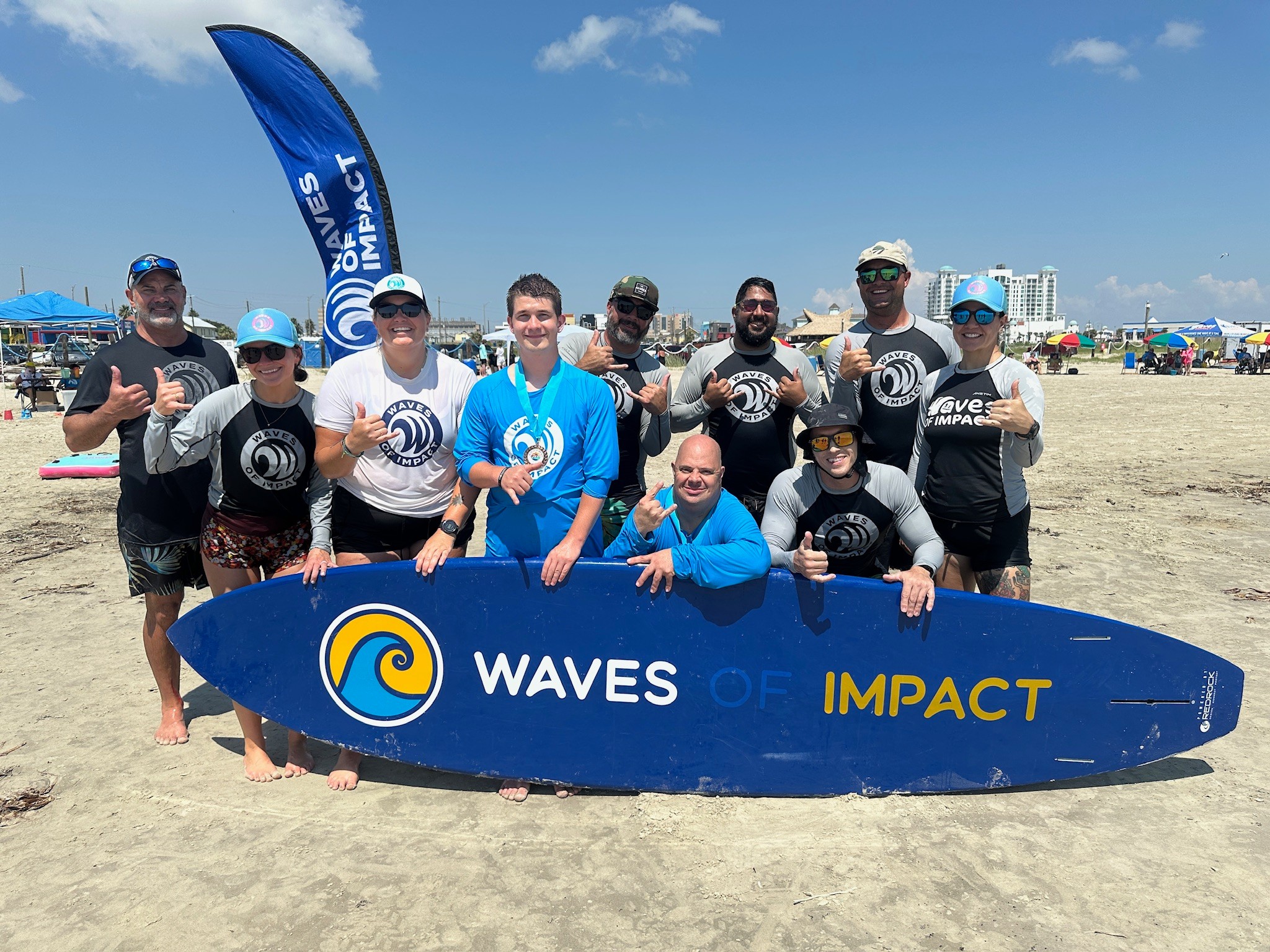 Group of people on the beach holding a large surfboard with 'WAVES IMPACT' written on it, blue sky above.