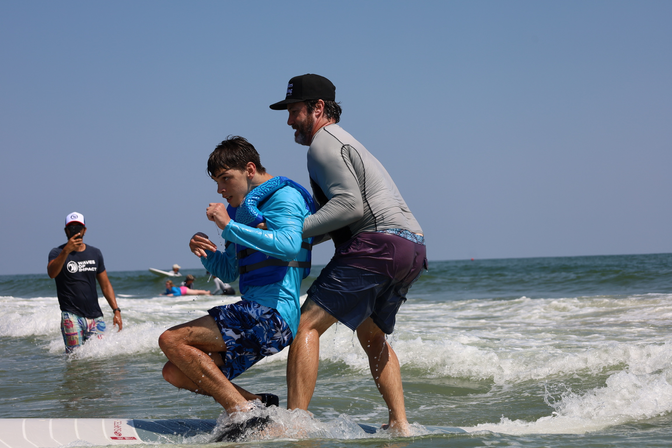 Two people playing in the ocean waves, one helping the other stand, with two more people in the background, under a clear sky.