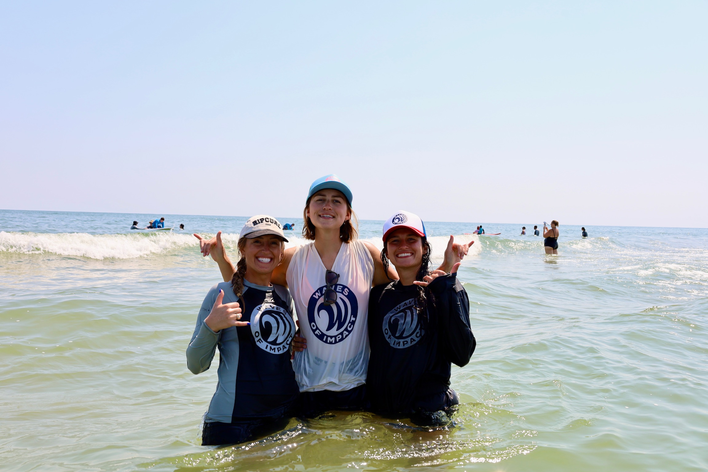 Three people standing in the ocean with waves, smiling, wearing hats and casual clothing, sunny day, distant boats and people in background.