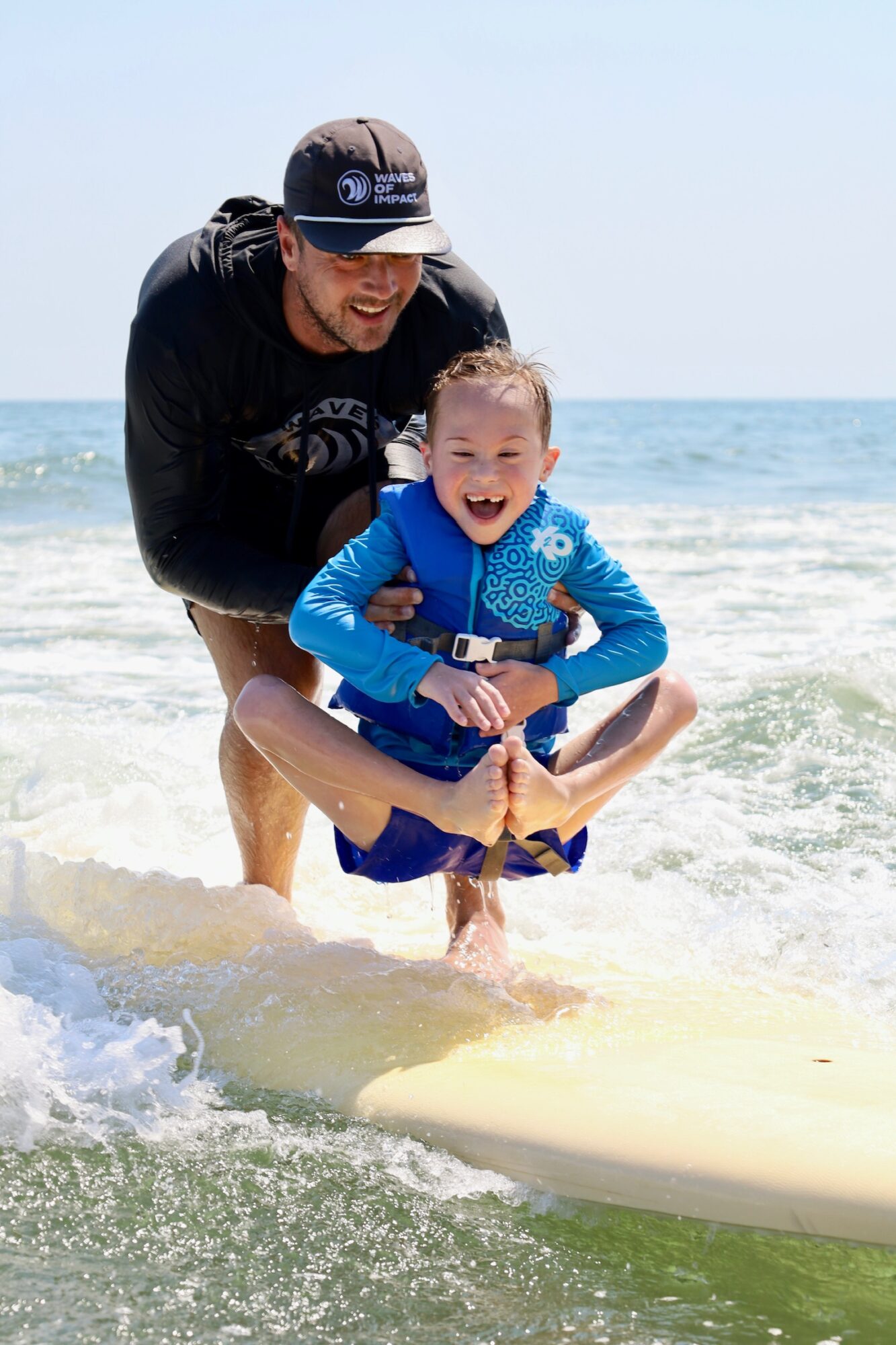 Adult and child smiling and playing in the ocean with a surfboard, sunny sky, and waves.