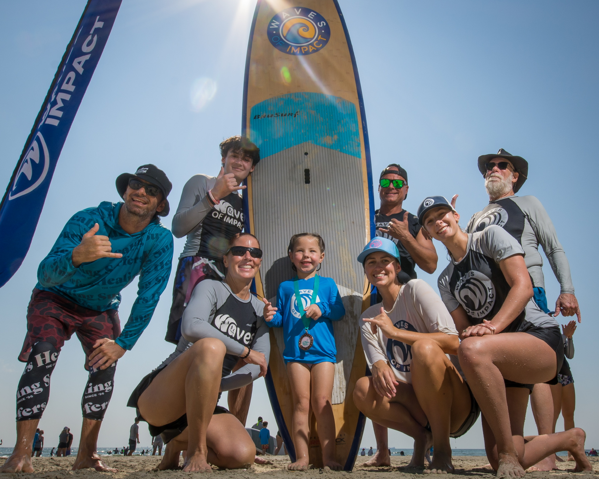 Group of people, including children, posing in front of a large paddleboard on a sandy beach under a clear sky.