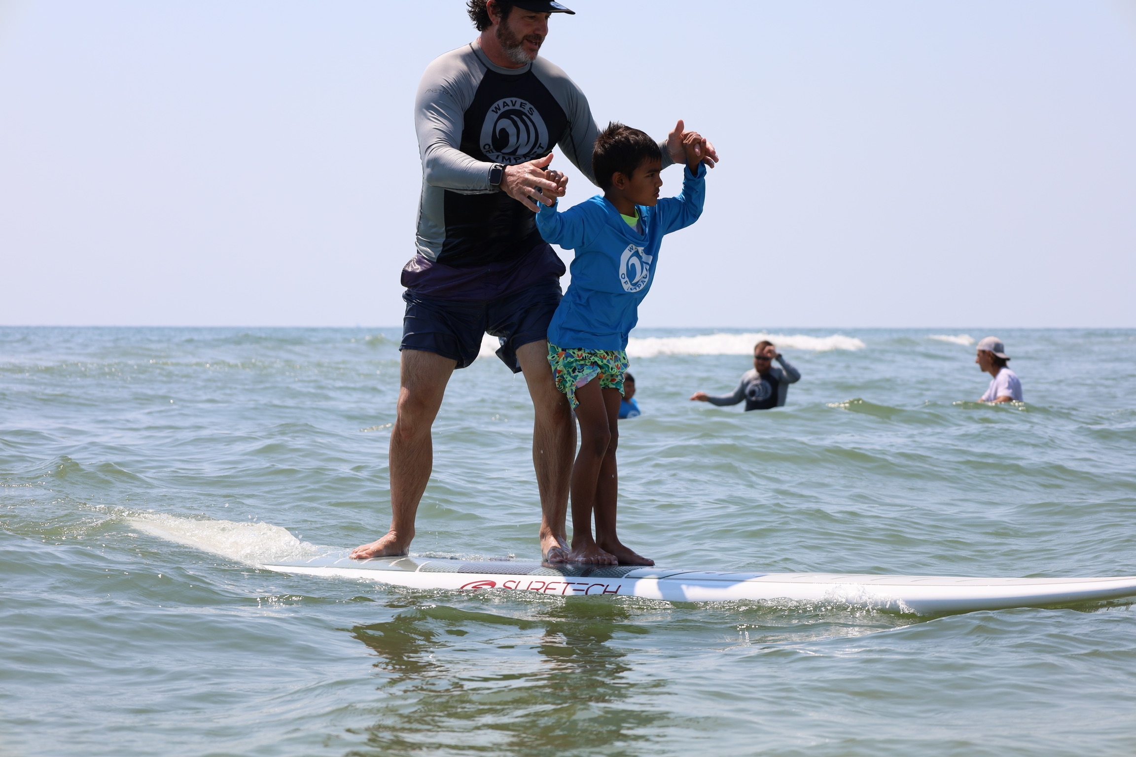 Adult and child stand on a surfboard in the ocean, holding hands, with other people swimming in the background.