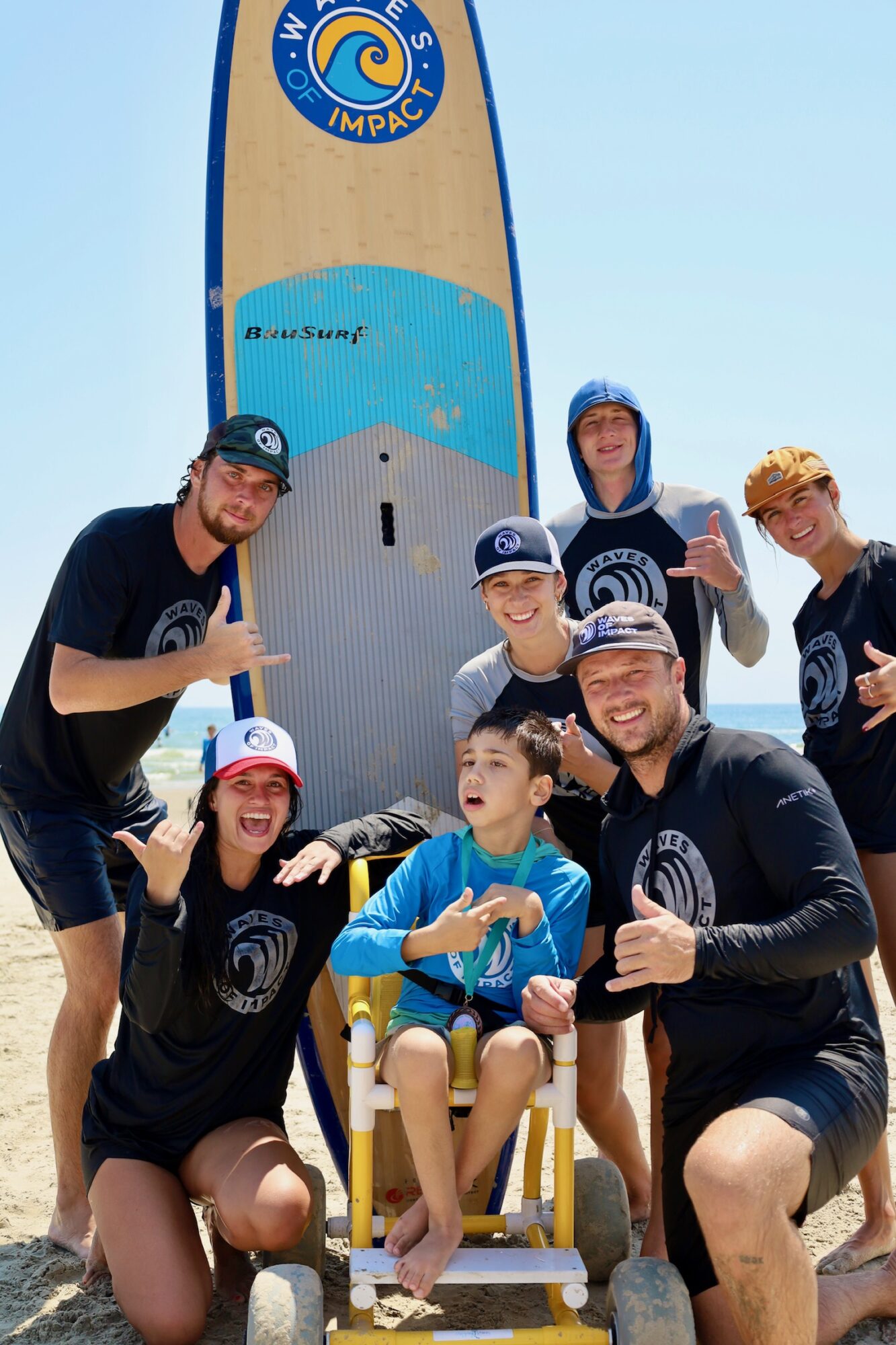 Group of people, including children, posing outdoors with a large paddleboard, some wearing matching shirts and hats, smiling and giving thumbs up.