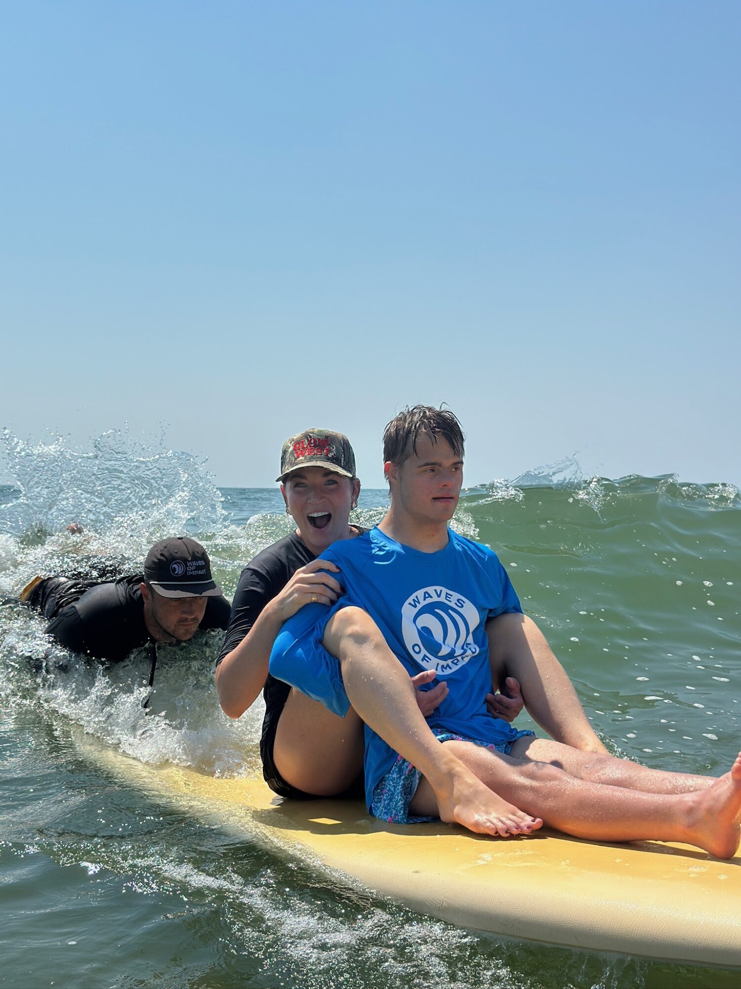 Three people sitting on a surfboard in the ocean, with waves and a clear blue sky in the background.
