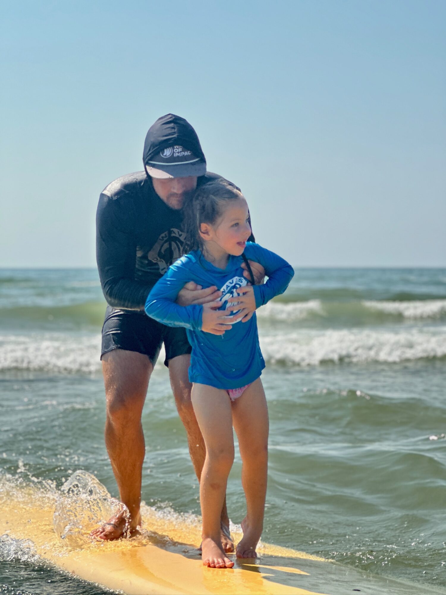 Adult and child standing on a surfboard in the ocean, with waves and sky in the background.