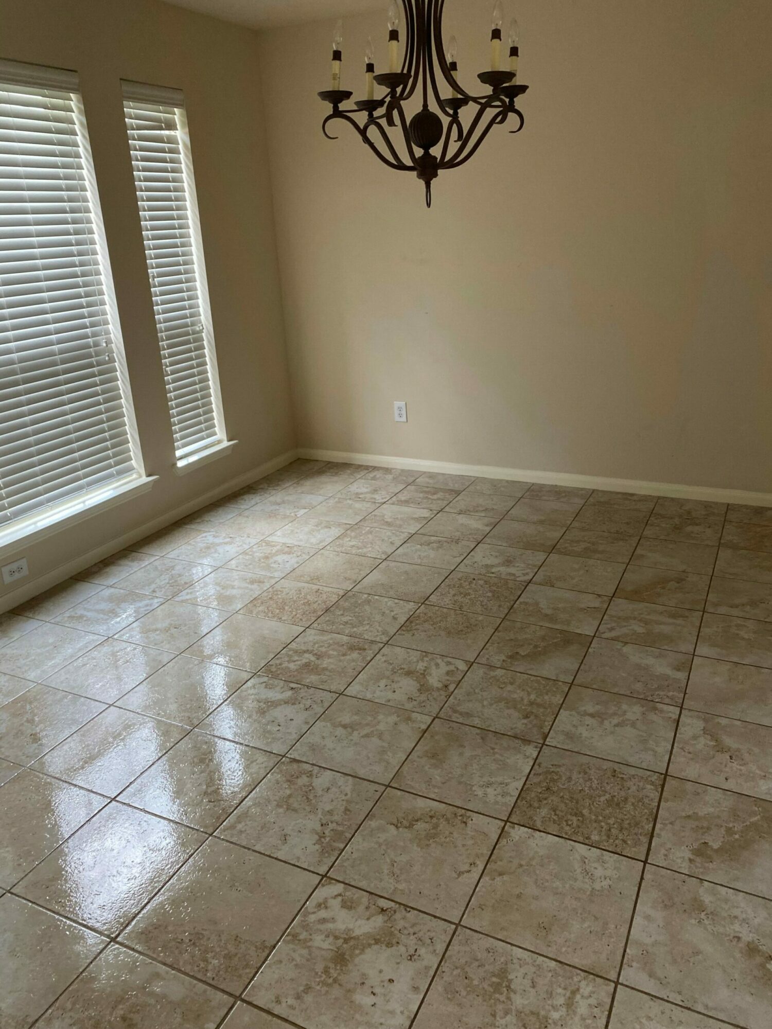 Empty room with tiled floor, beige walls, and a chandelier, sunlight coming through window blinds.