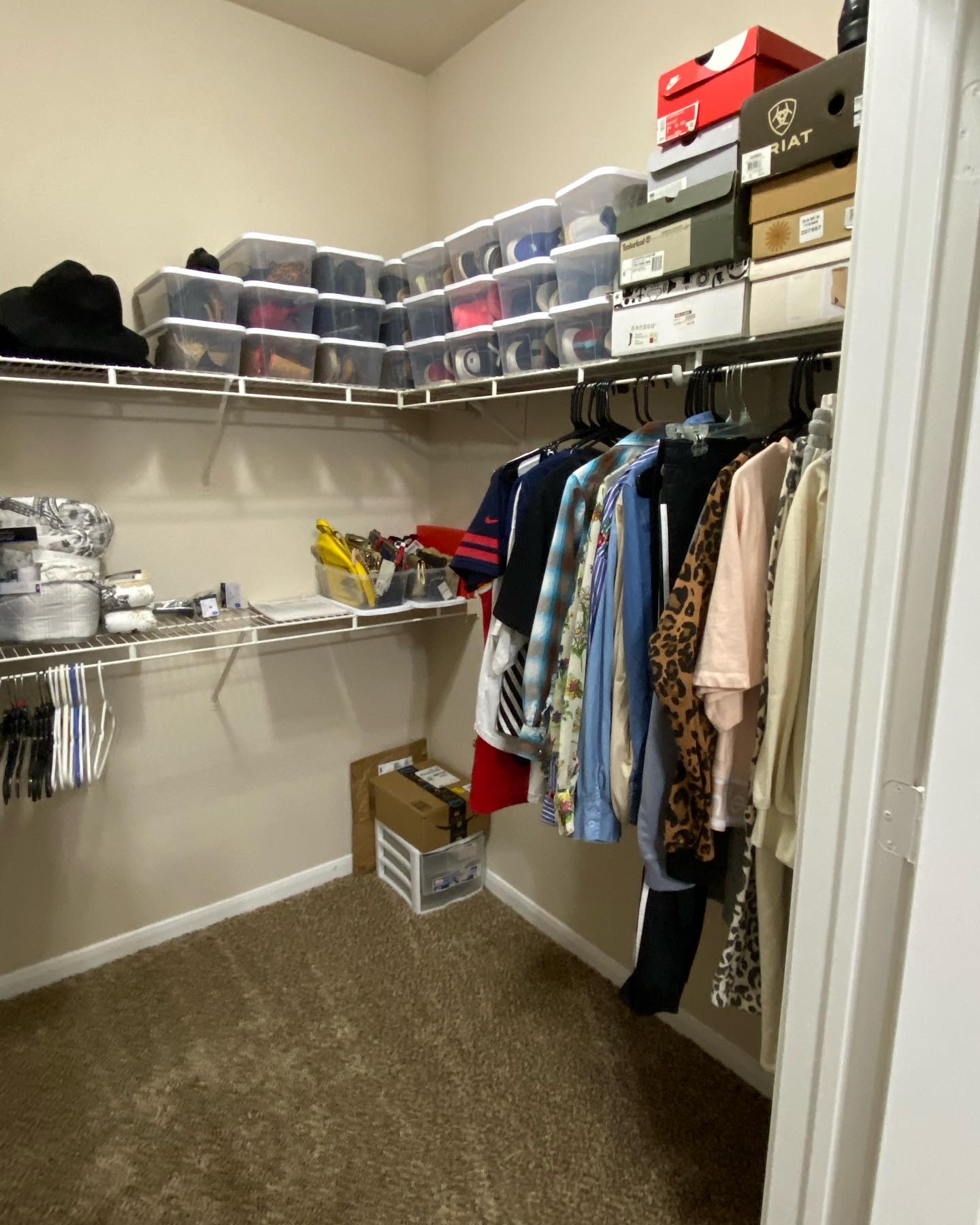 Closet with shelves of shoe boxes, hanging clothes, and storage boxes on top, with a carpeted floor.