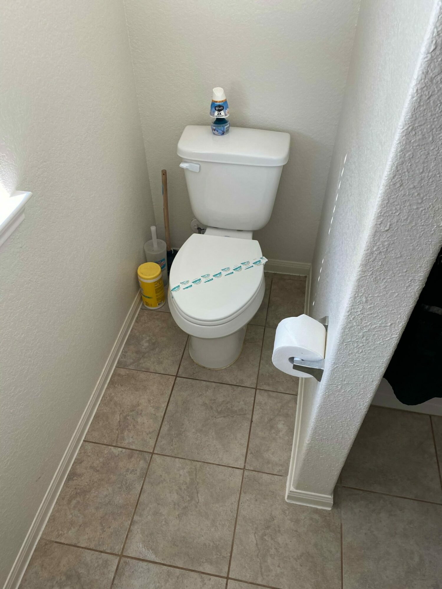 Toilet with a closed lid, a yellow cleaning bottle, and a toilet paper roll on a wall-mounted holder, in a bathroom corner.