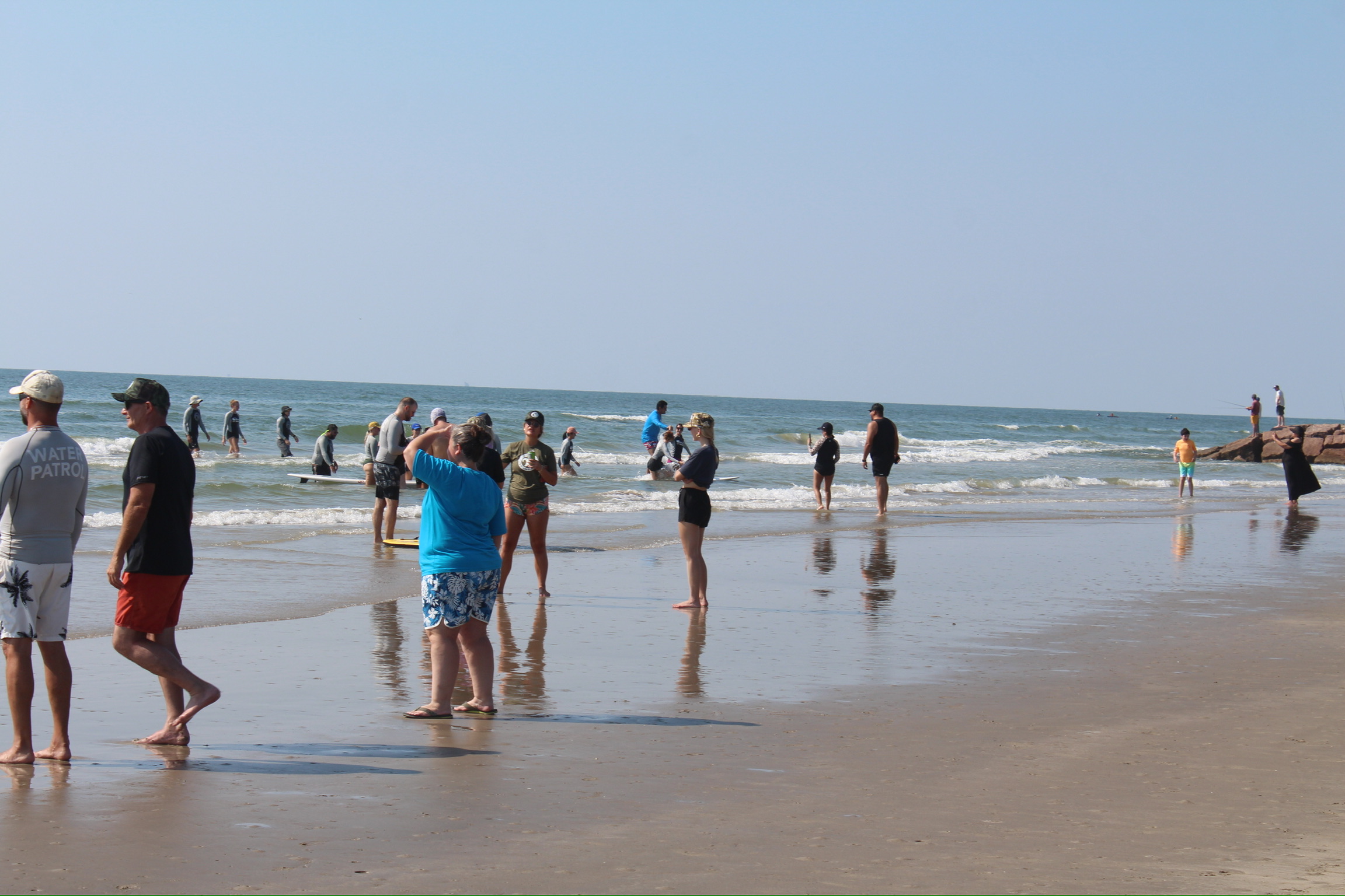 People walking and standing on a sandy beach near the ocean, some in the water, under a clear sky.
