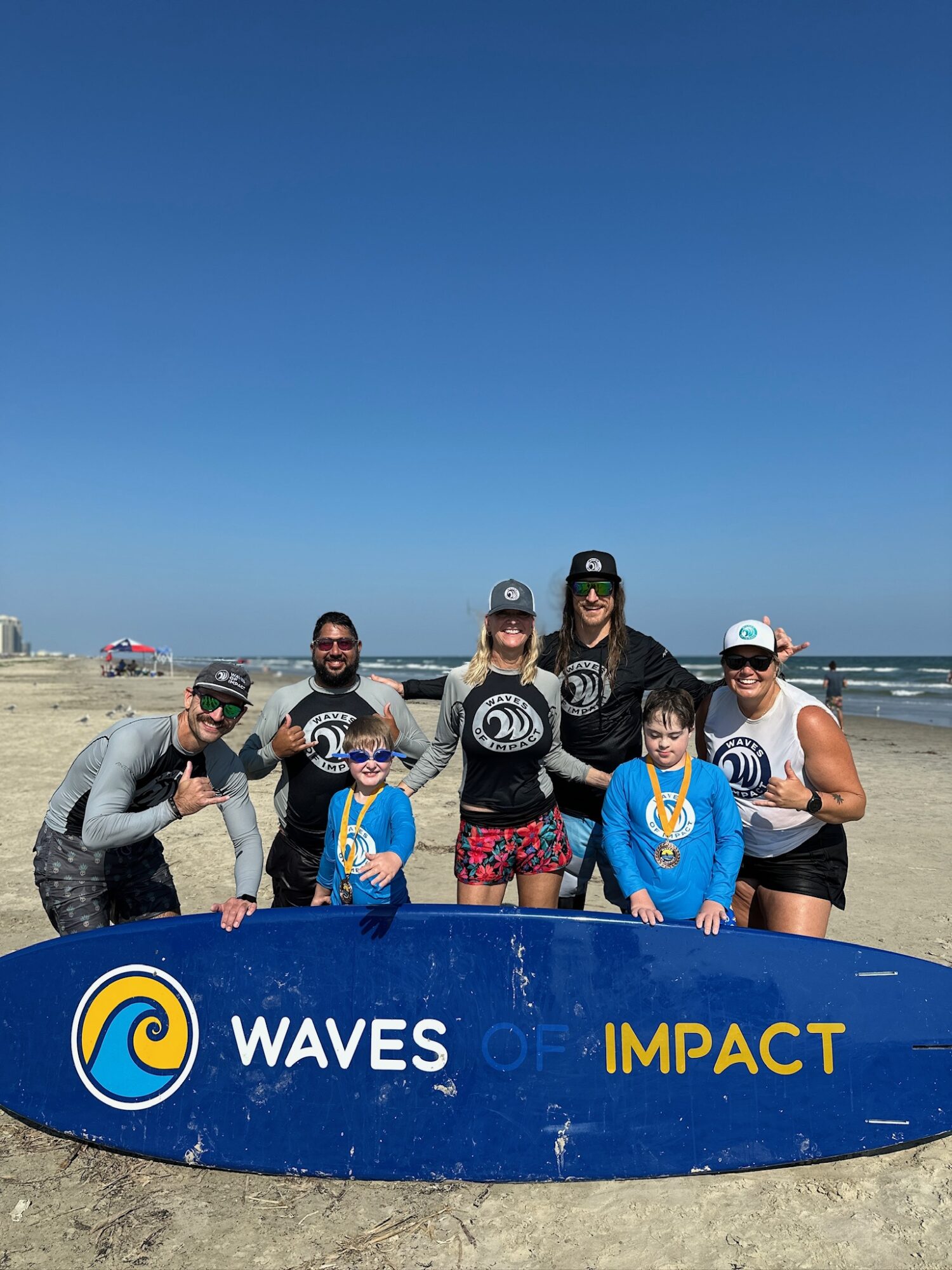 Group of people on the beach holding a blue and yellow sign with waves and impact logos, under a clear blue sky.
