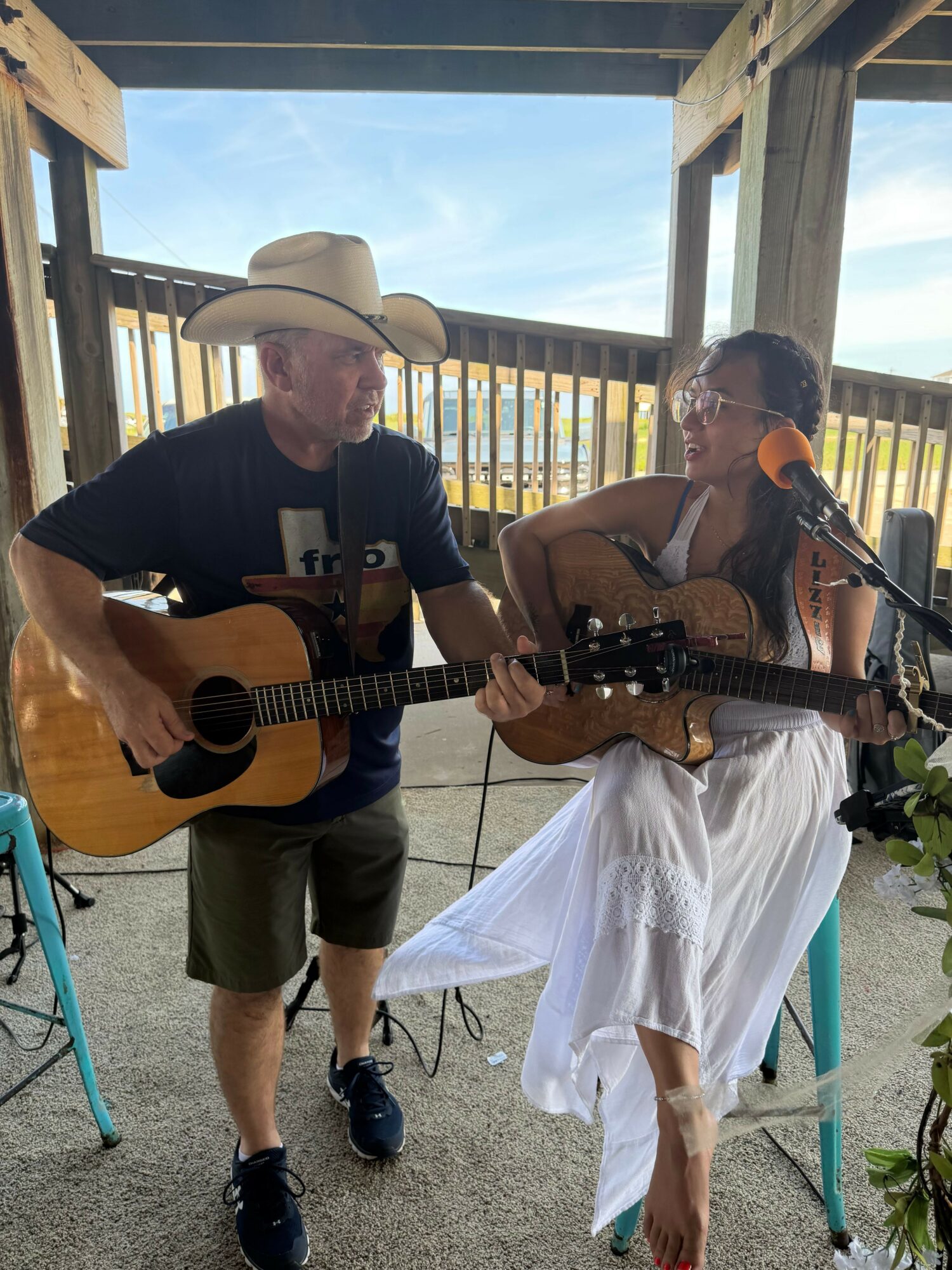 Two people, one playing guitar and the other singing, on a balcony with a wooden railing and blue sky background.