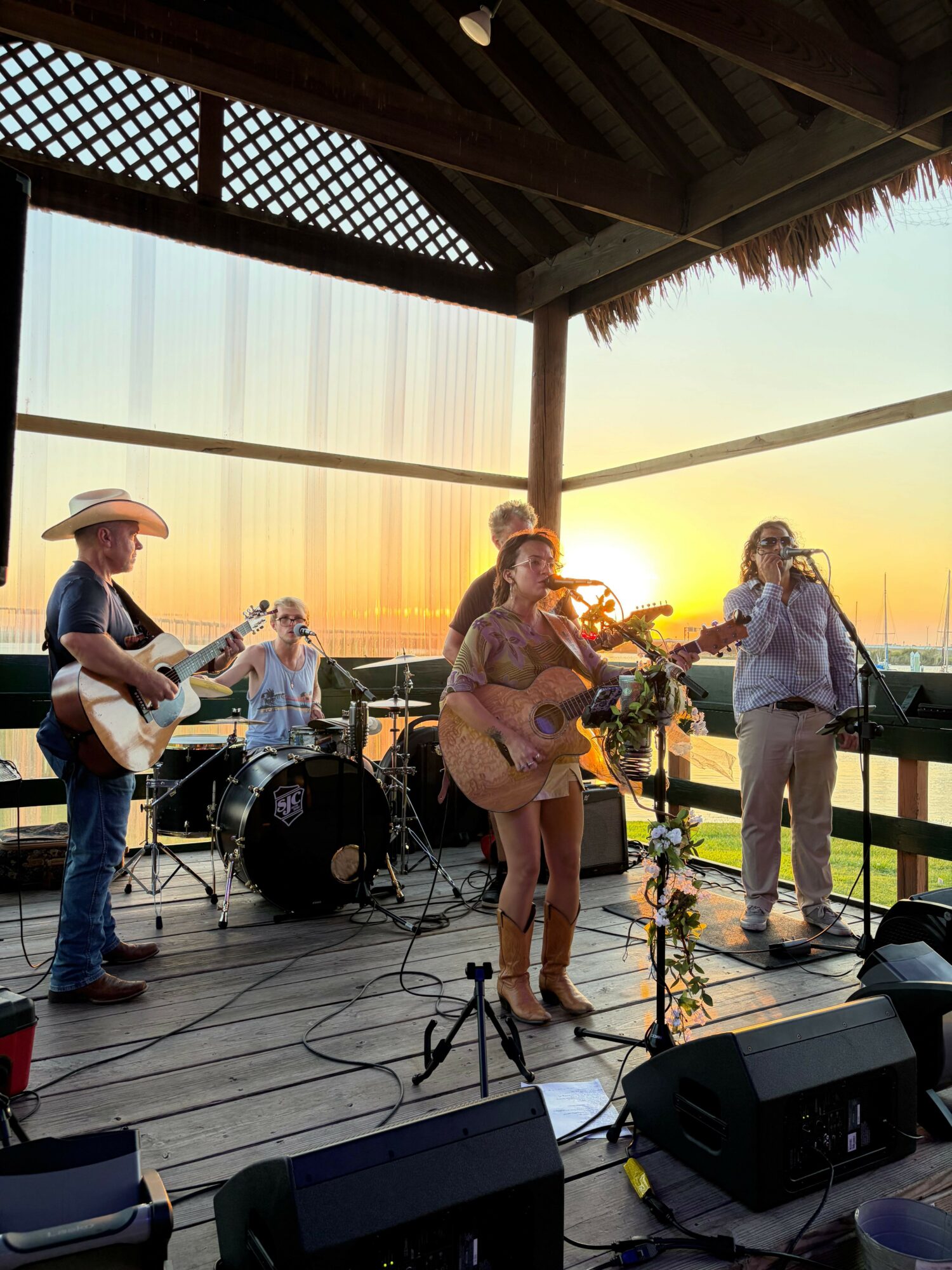 Two women and a man on a wooden deck during sunset, with musical equipment and a woman in the background.