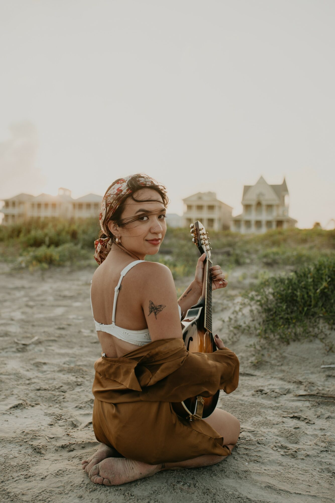 Young woman sitting on ground holding a guitar, with houses in the background, outdoors during daytime.