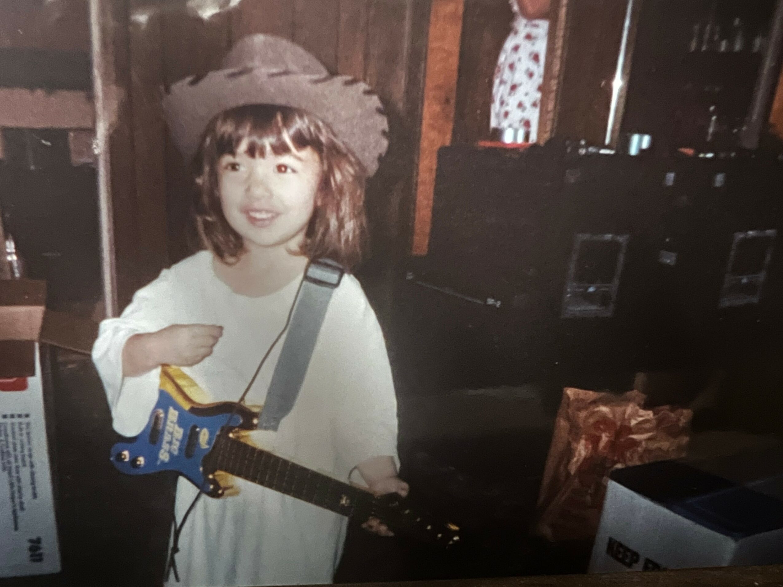 Child with a guitar and a hat, smiling, indoors with wooden walls and equipment in background.