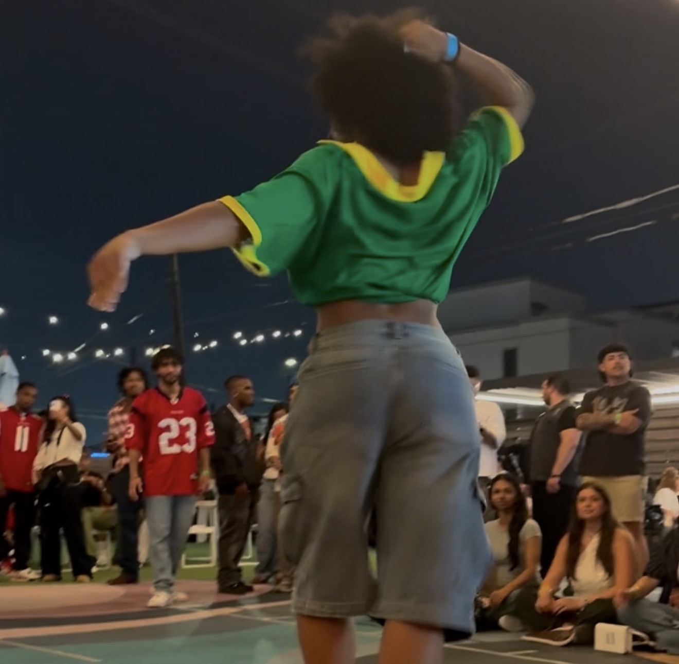 Person with curly hair wearing a green and yellow sports jersey and gray shorts, standing outdoors at night with a group of people around.