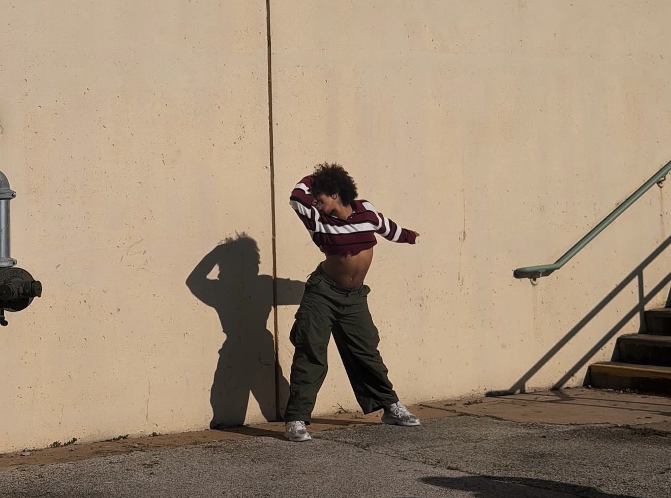 Person with curly hair lifting shirt to reveal stomach, standing against a beige wall with shadow, stairs on right.