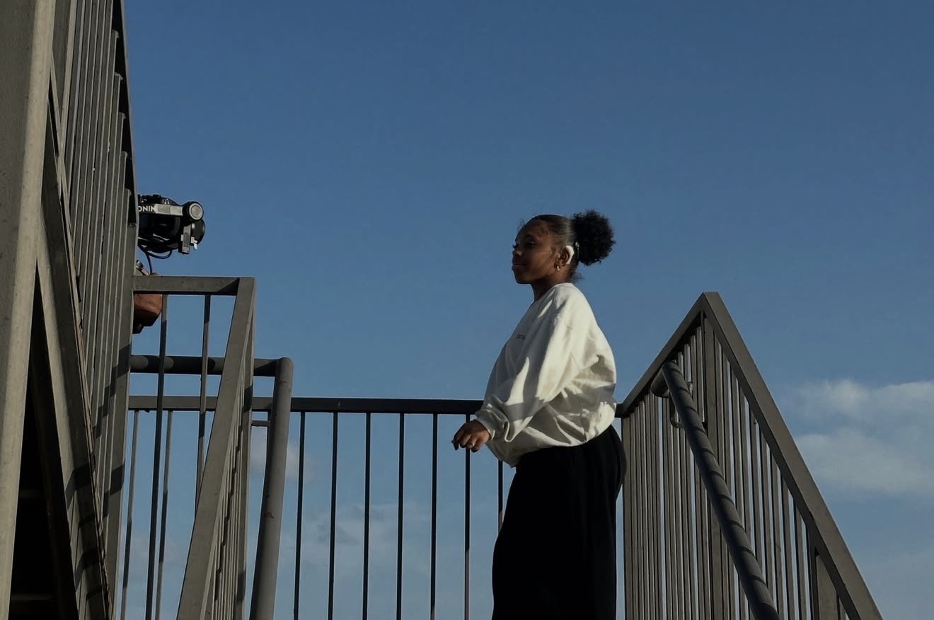 Young woman standing on a staircase railing against a blue sky, wearing a white top and black pants.