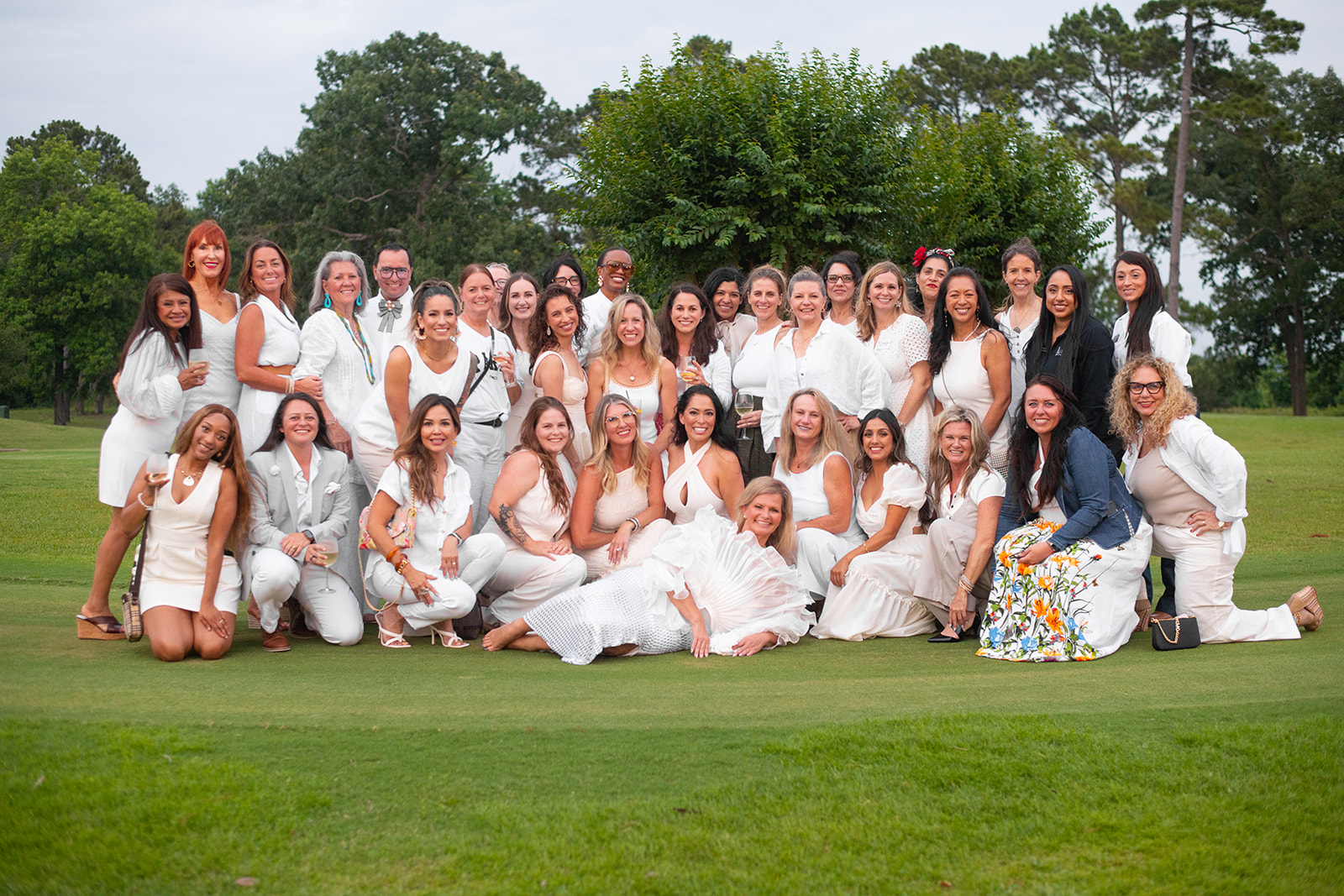 Group of women in white dresses outdoors on grass, with trees and sky in background.