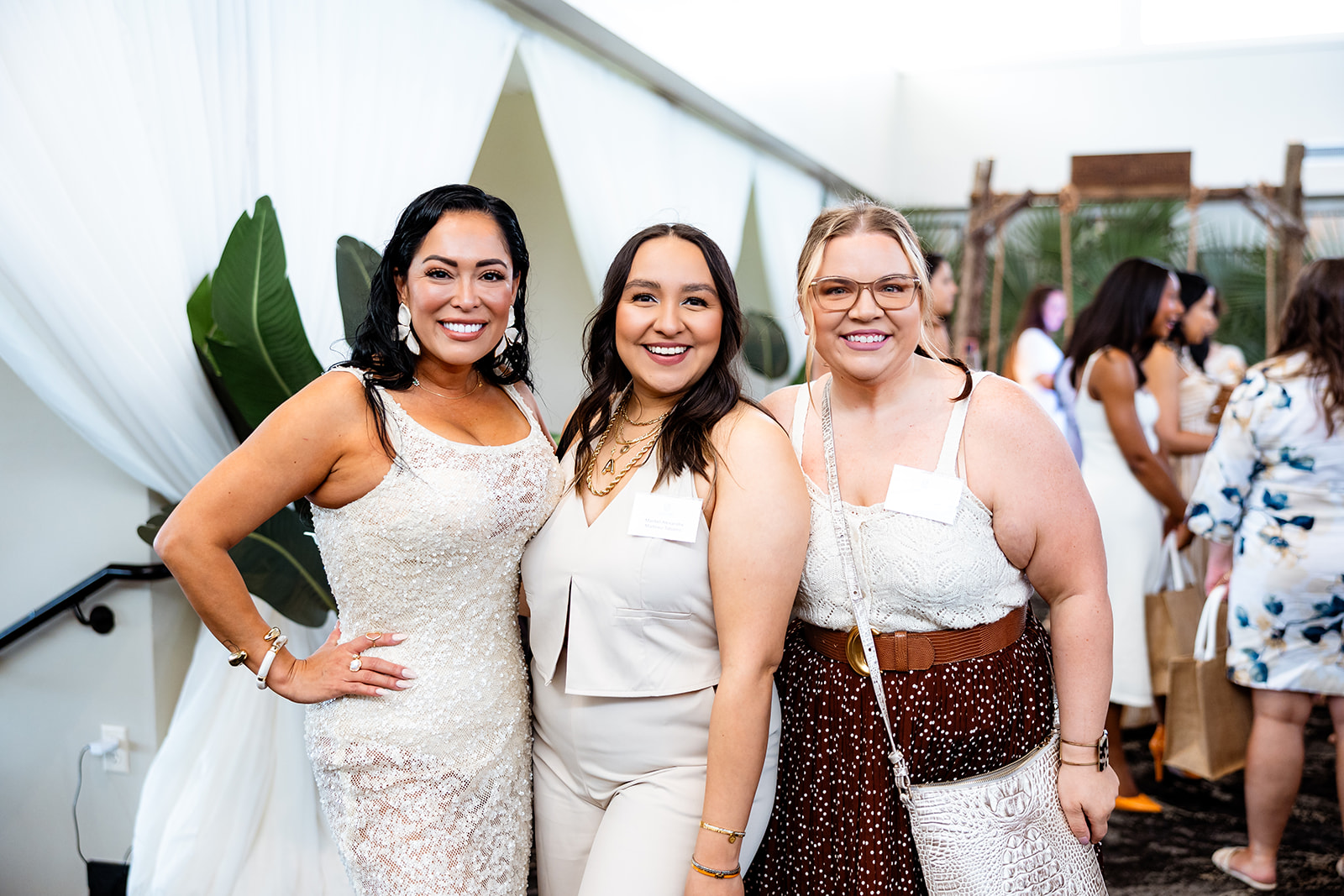 Three women smiling and posing together indoors, dressed in light-colored outfits, with a large plant and other people in the background.