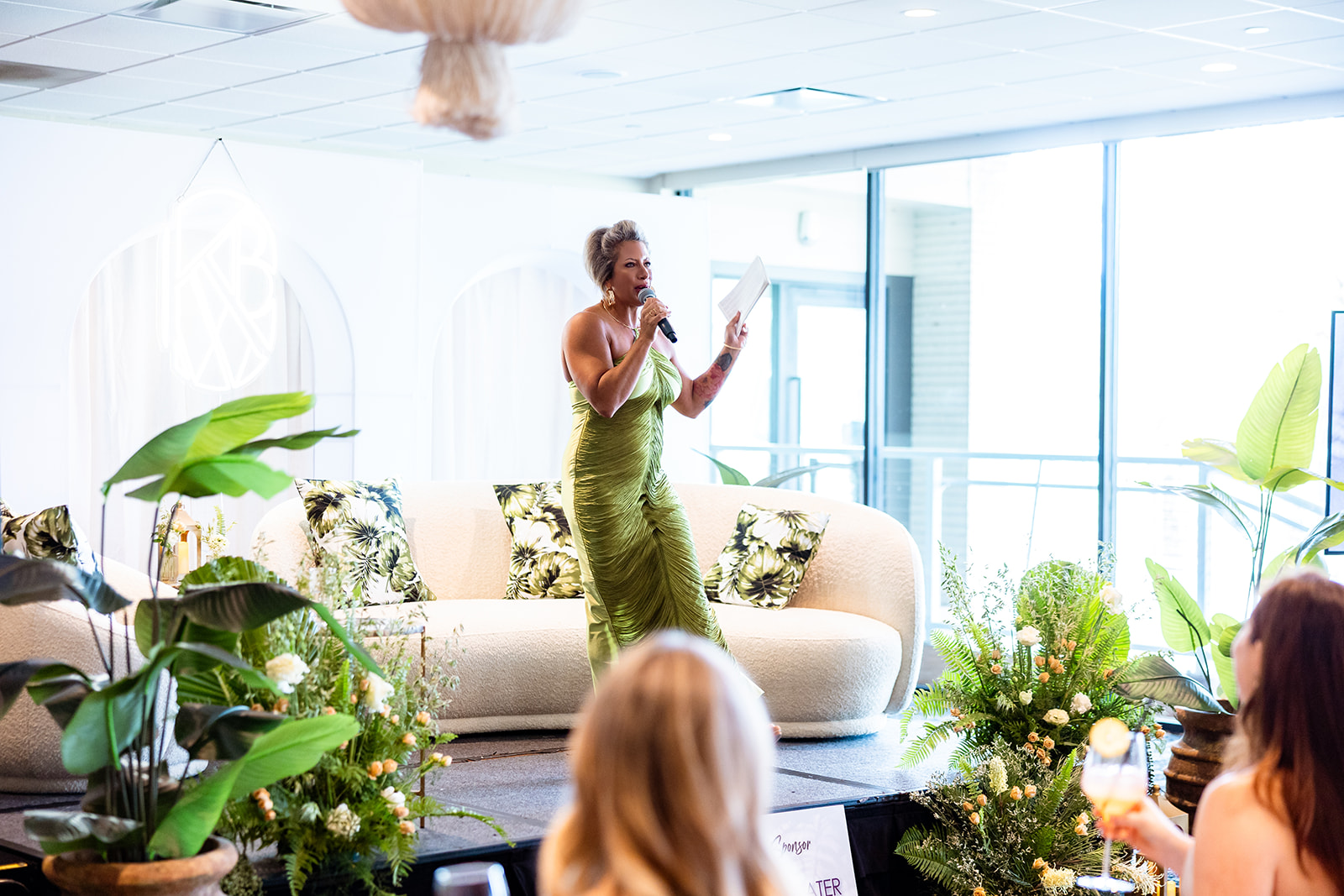 Woman in green dress speaking on stage with plants and a sofa, audience visible in foreground, bright room with large windows.